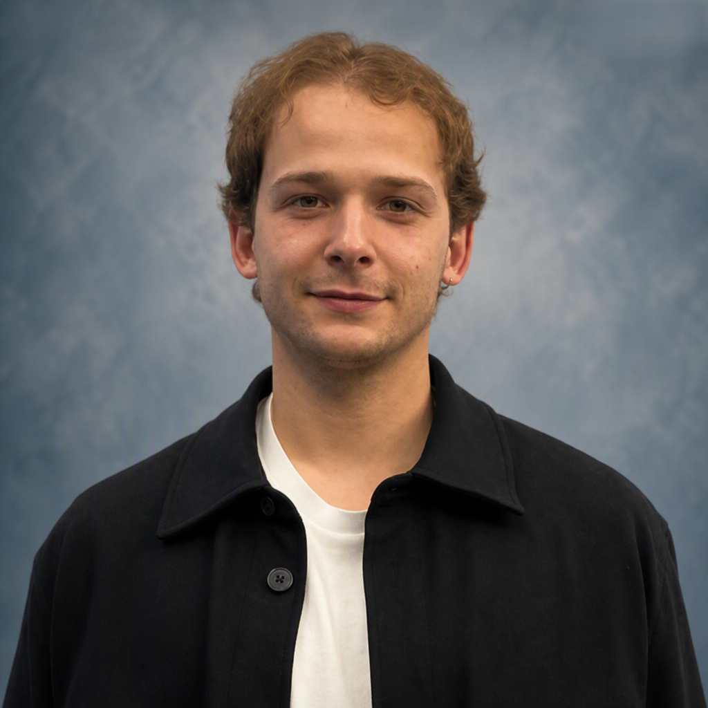 Portrait of a young man with light brown curly hair, wearing a black jacket over a white t-shirt, standing against a blue-gray background.