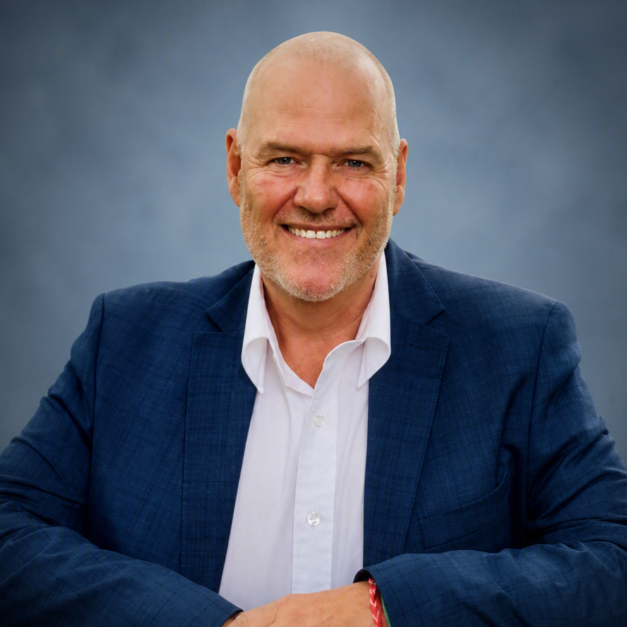 A bald man with a beard wearing a white dress shirt and a dark blue blazer, smiling against a gray background.