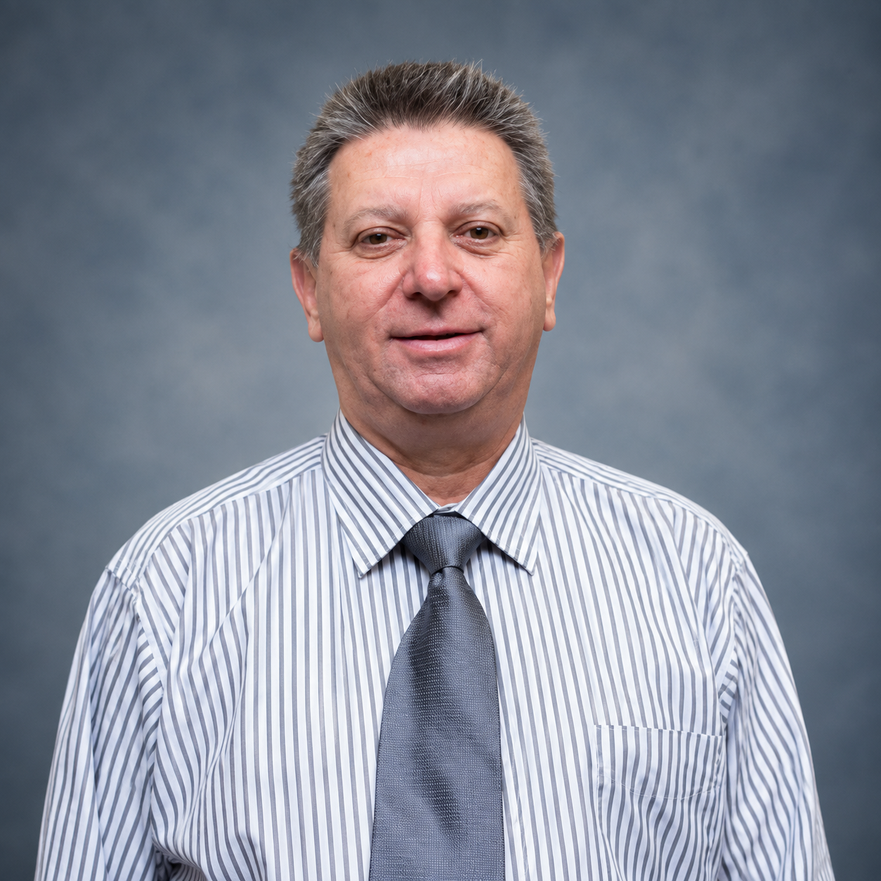 A middle-aged man with gray hair, wearing a striped dress shirt and a gray tie, standing against a plain gray background.