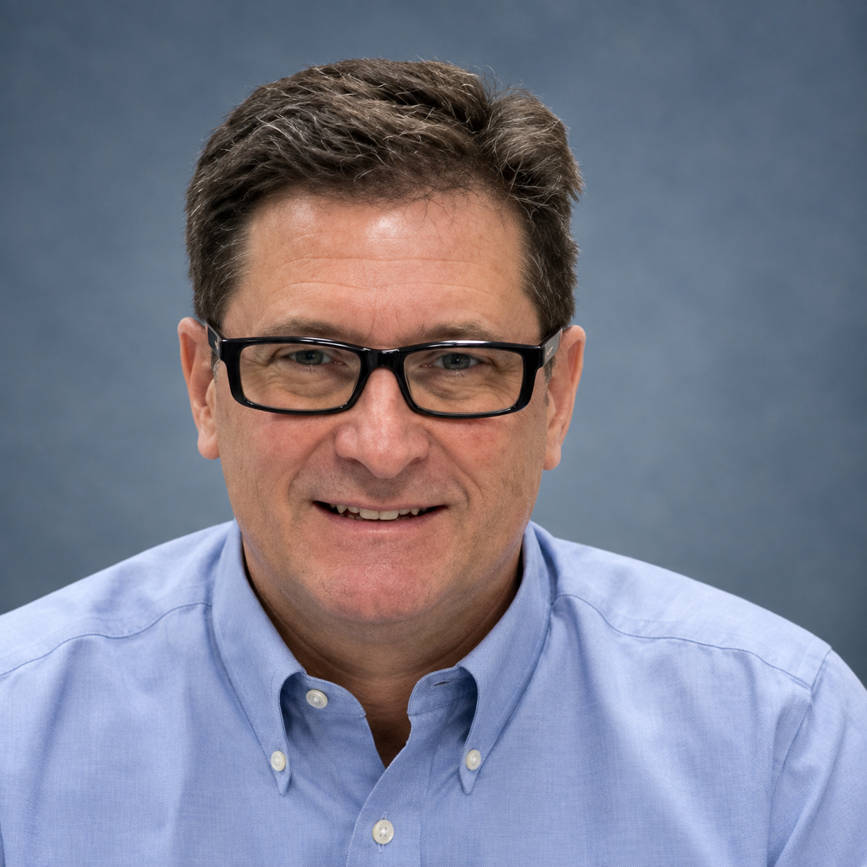 A middle-aged man with dark hair, glasses, and light skin, smiling, wearing a light blue collared shirt against a plain gray background.