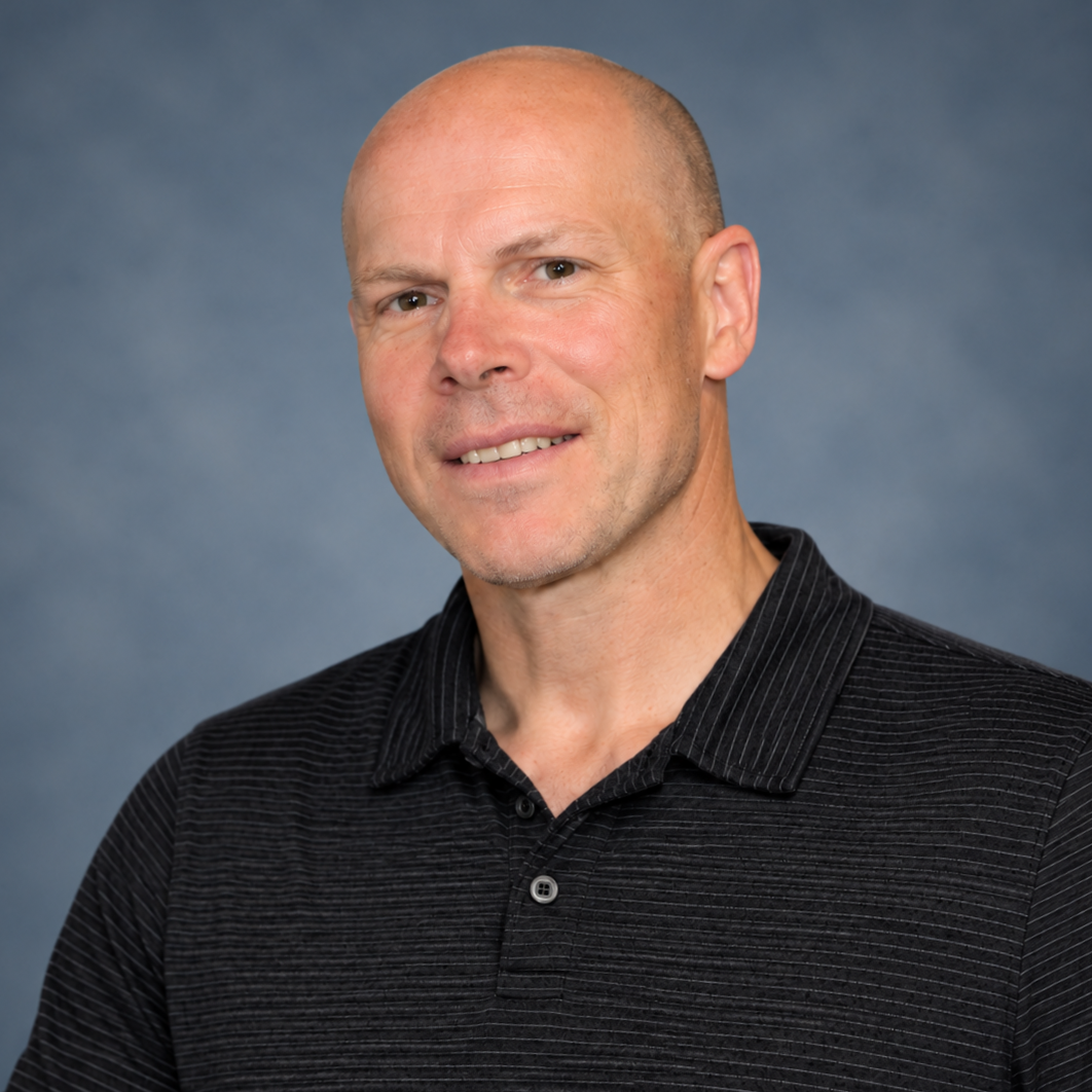 A middle-aged man with a shaved head and light skin, wearing a black collared shirt, smiling slightly against a plain, dark blue background.