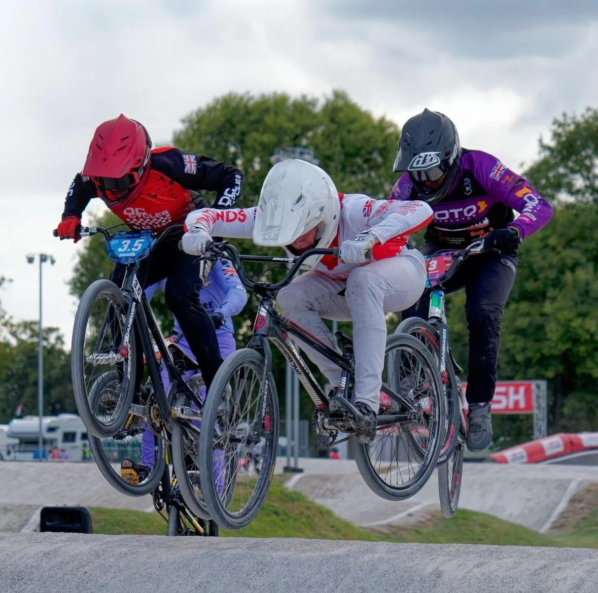 Three BMX riders in gear jumping through an obstacle on a BMX track, with trees and cloudy sky in the background.