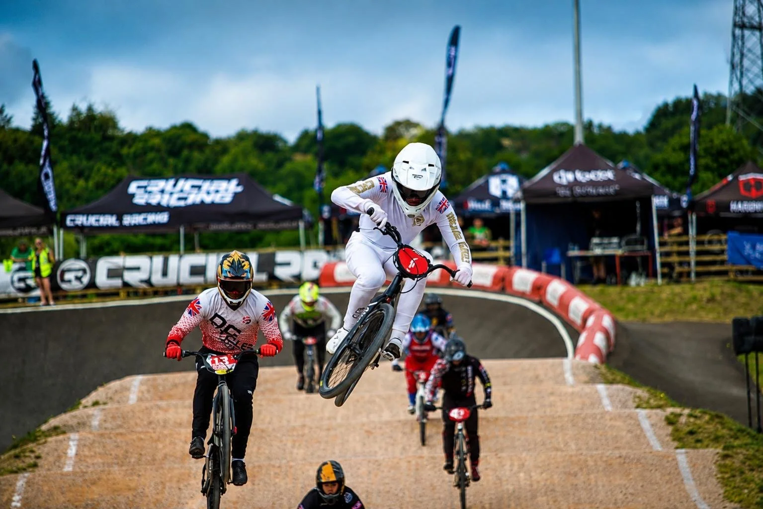 BMX riders in a race, one in mid-air performing a jump, others riding along a dirt track with race banners and tents in the background.