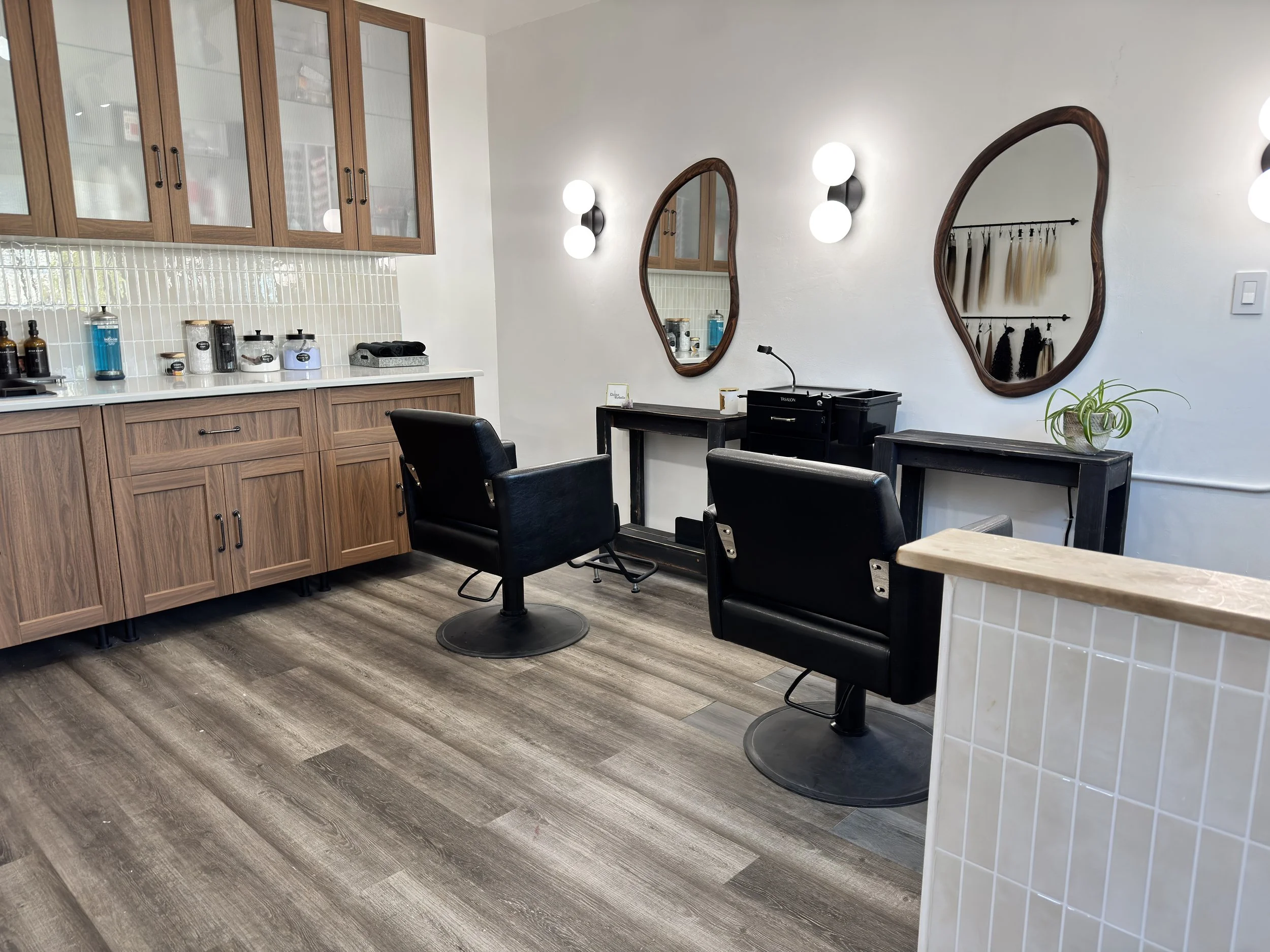 Interior of a modern hair salon with wooden cabinets, black salon chairs, two oval mirrors with wooden frames, wall-mounted light fixtures with globe bulbs, a small table with a potted plant, and hair color swatches hanging on a rack.