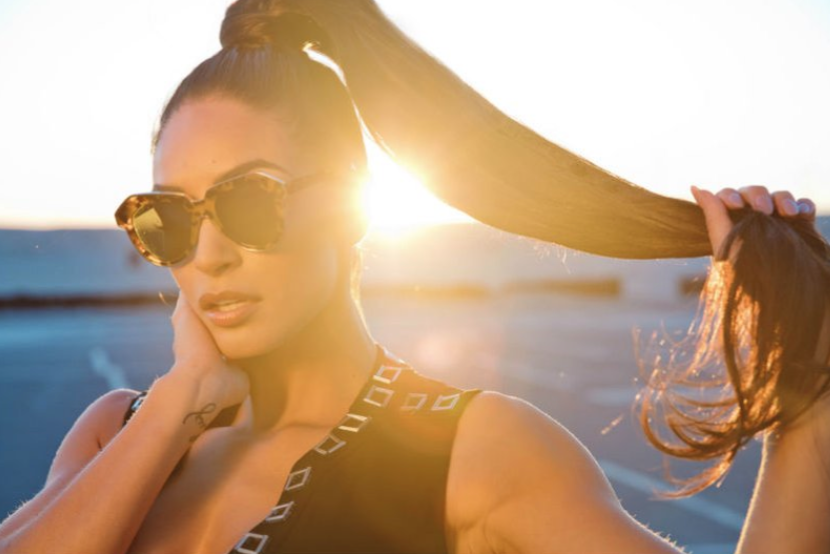Woman with long hair wearing sunglasses, holding her hair, with sunlight in the background.