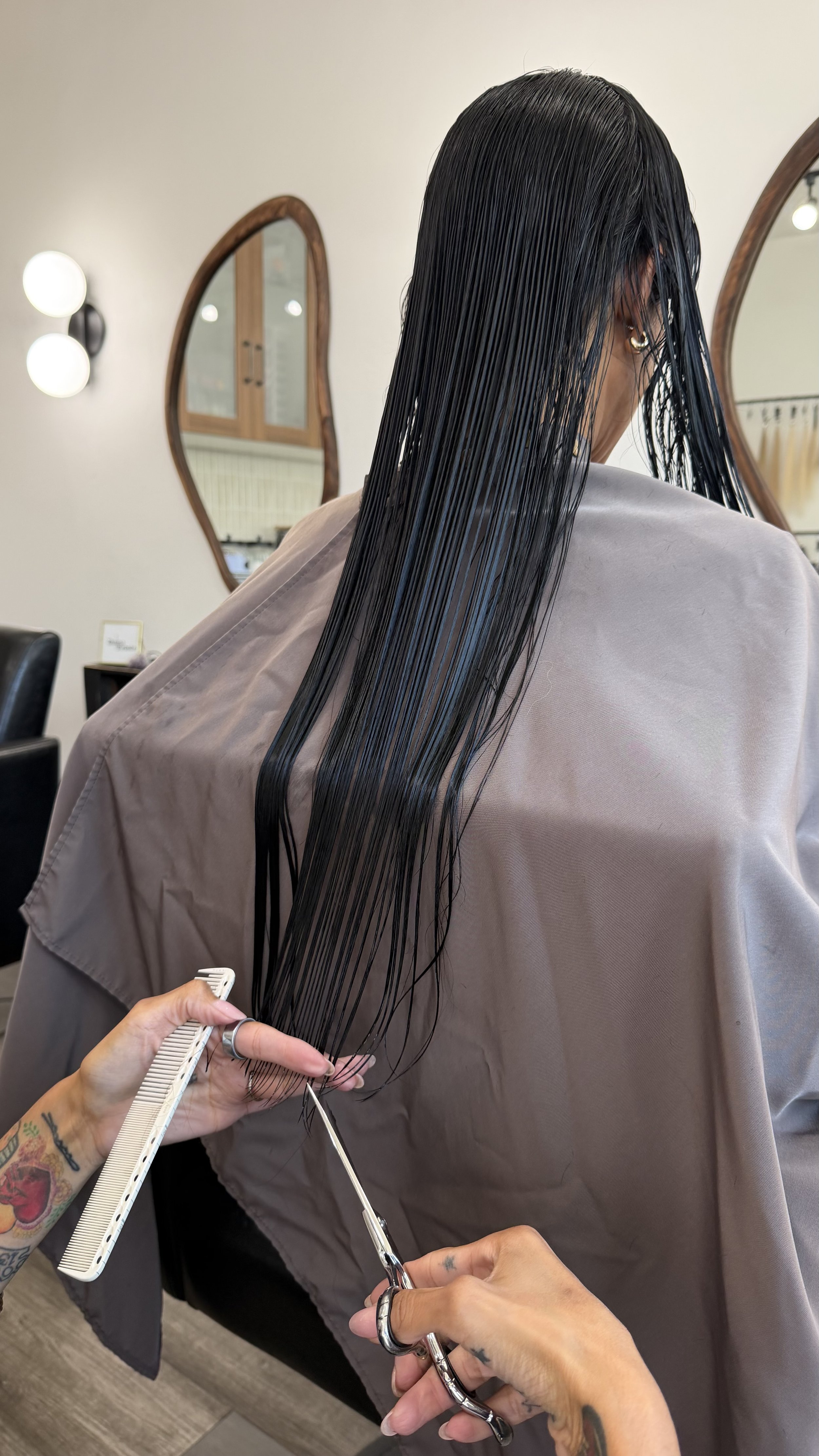 A person with dark, wet hair sitting in a salon chair, getting hair cut or styled. A hairstylist is holding scissors and a comb.