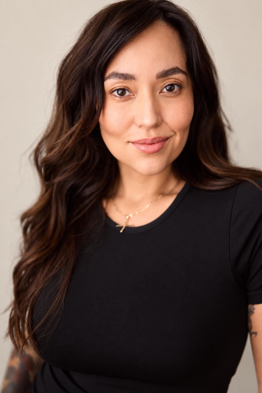 Close-up portrait of a young woman with long, wavy brown hair wearing a black shirt and a gold cross necklace, smiling slightly.