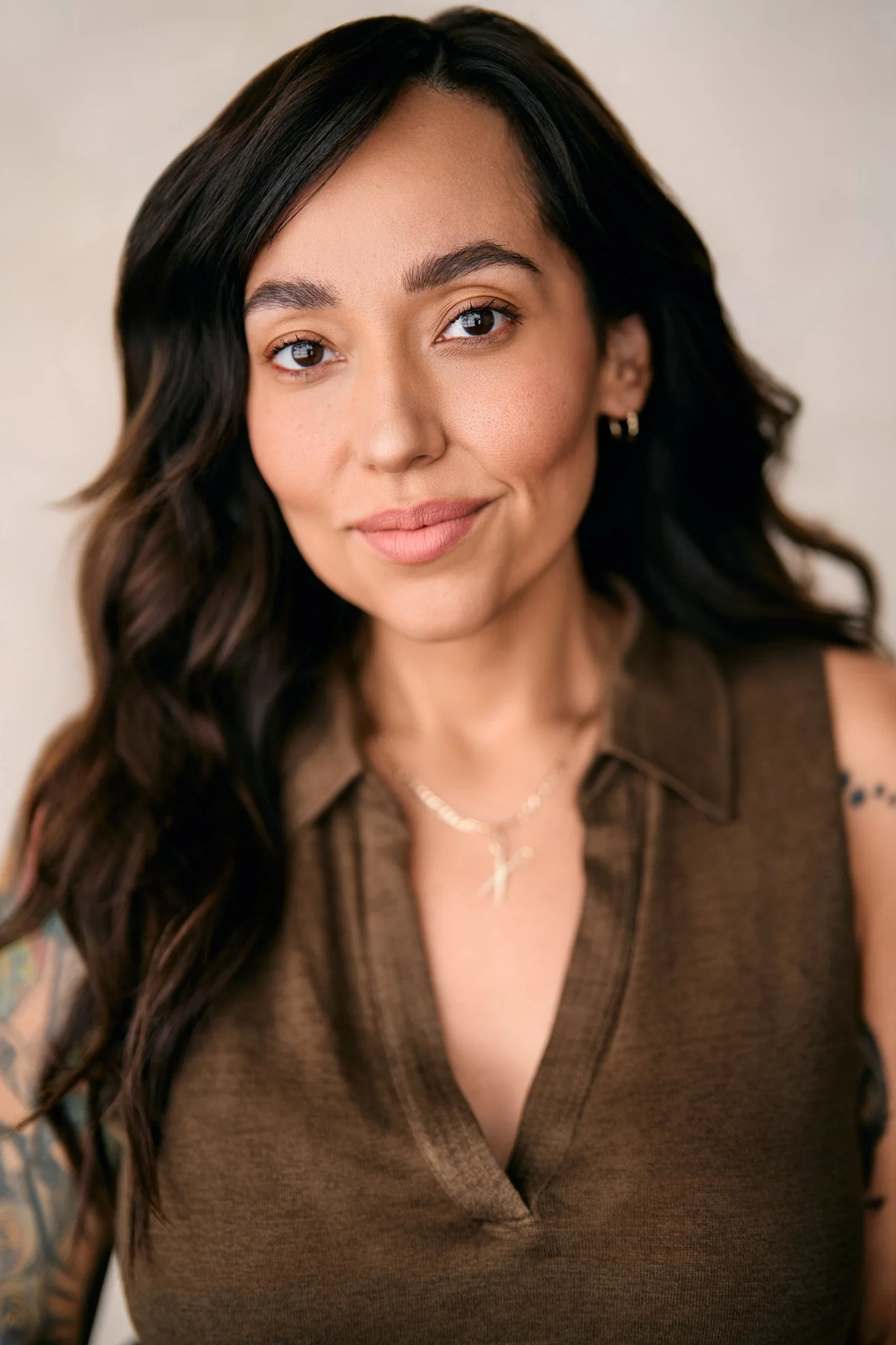A woman with long, dark wavy hair wearing a brown top, gold earrings, and a gold necklace, smiling softly at the camera against a neutral background.
