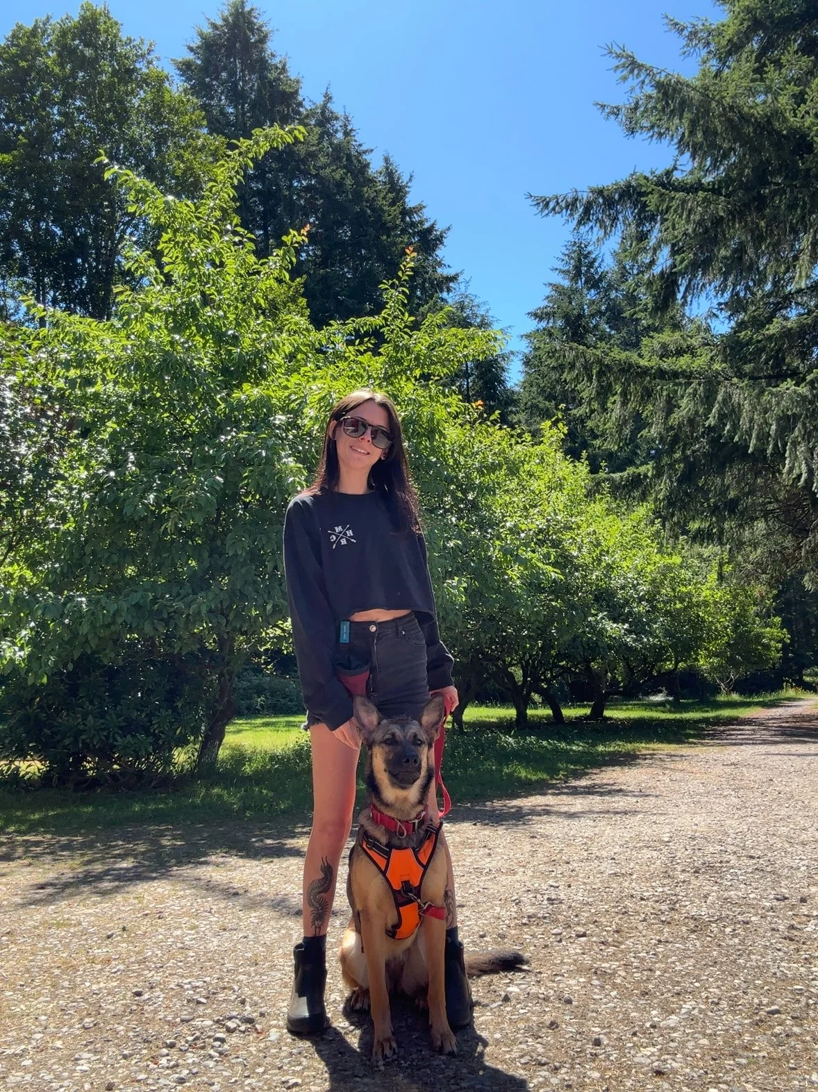 A woman wearing sunglasses and a dark long-sleeved top standing outdoors on a sunny day with her German Shepherd dog wearing an orange harness. They are on a gravel path surrounded by green trees and foliage under a clear blue sky.