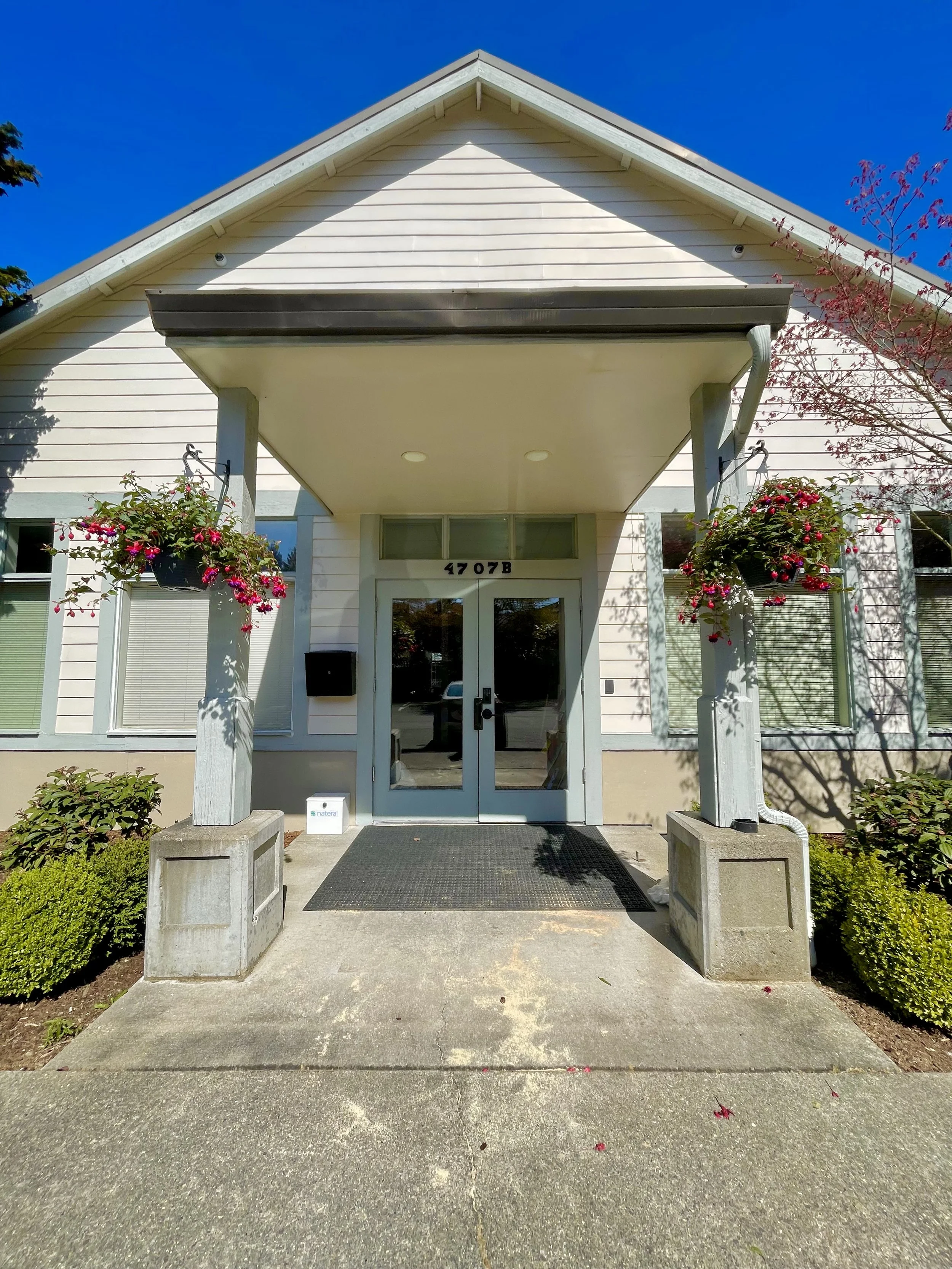 A welcoming front entrance of a pelvic floor physical therapy clinic featuring two hanging flower baskets and surrounded by bushes. The accessible building has a gabled roof and a concrete walkway from free parking leading to a glass door.