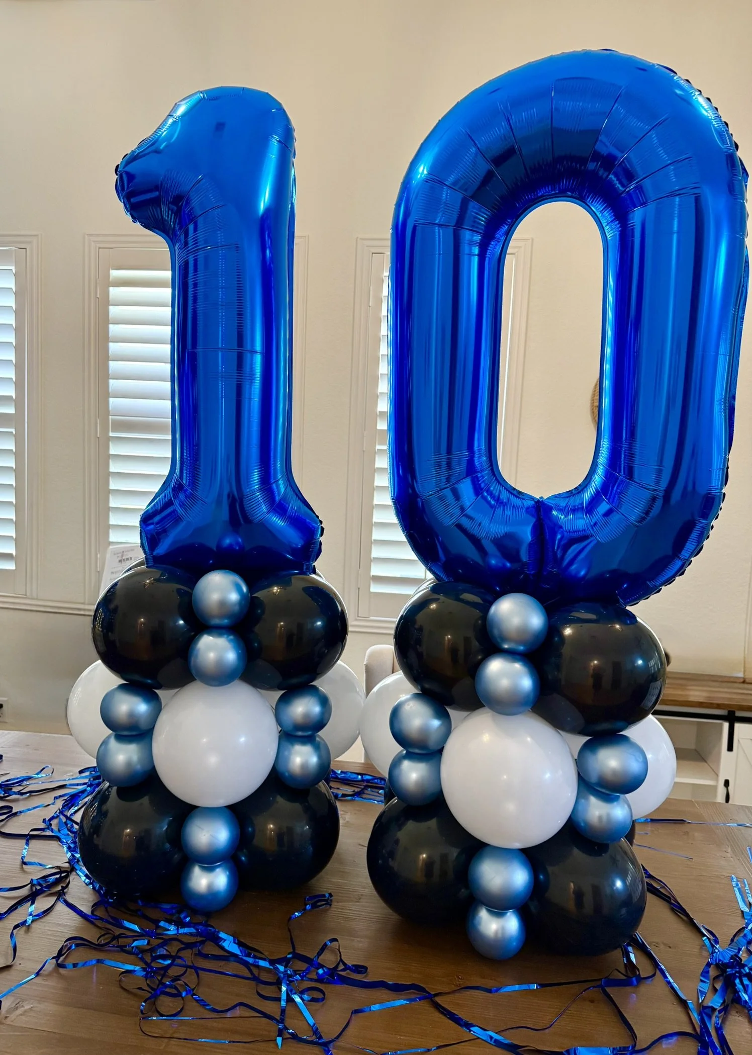 Large blue number balloons forming the number 10, with black, silver, and white balloons underneath, decorated with blue curly ribbons on a wooden floor in front of white window shutters.
