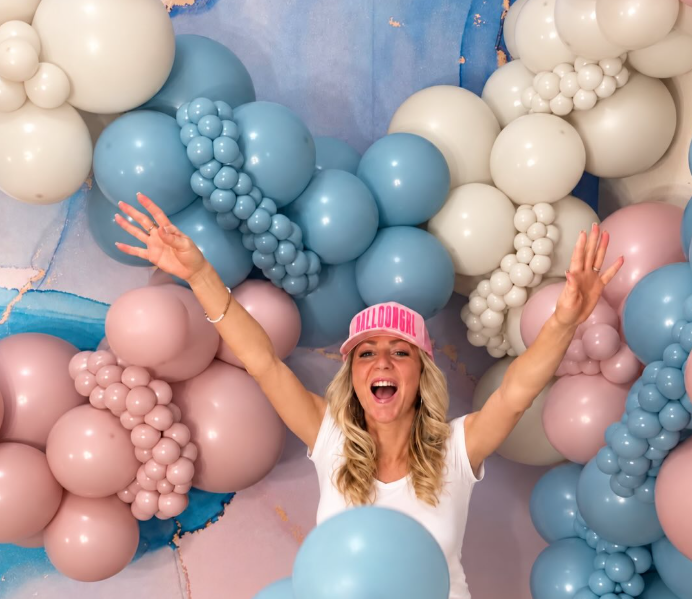 Woman celebrating with arms raised in front of a colorful balloon arch.