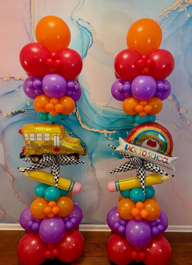 Colorful balloon columns with themes of back to school, featuring balloons in red, purple, orange, teal, and yellow, topped with school bus and rainbow-shaped balloons, set against a marbled blue wall.