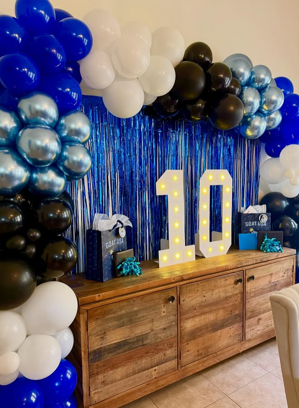 Balloon arch with white, black, blue, and silver balloons framing a backdrop of blue metallic fringe. Large lit up number ten on a wooden sideboard, with gift bags and party favors for a celebration.