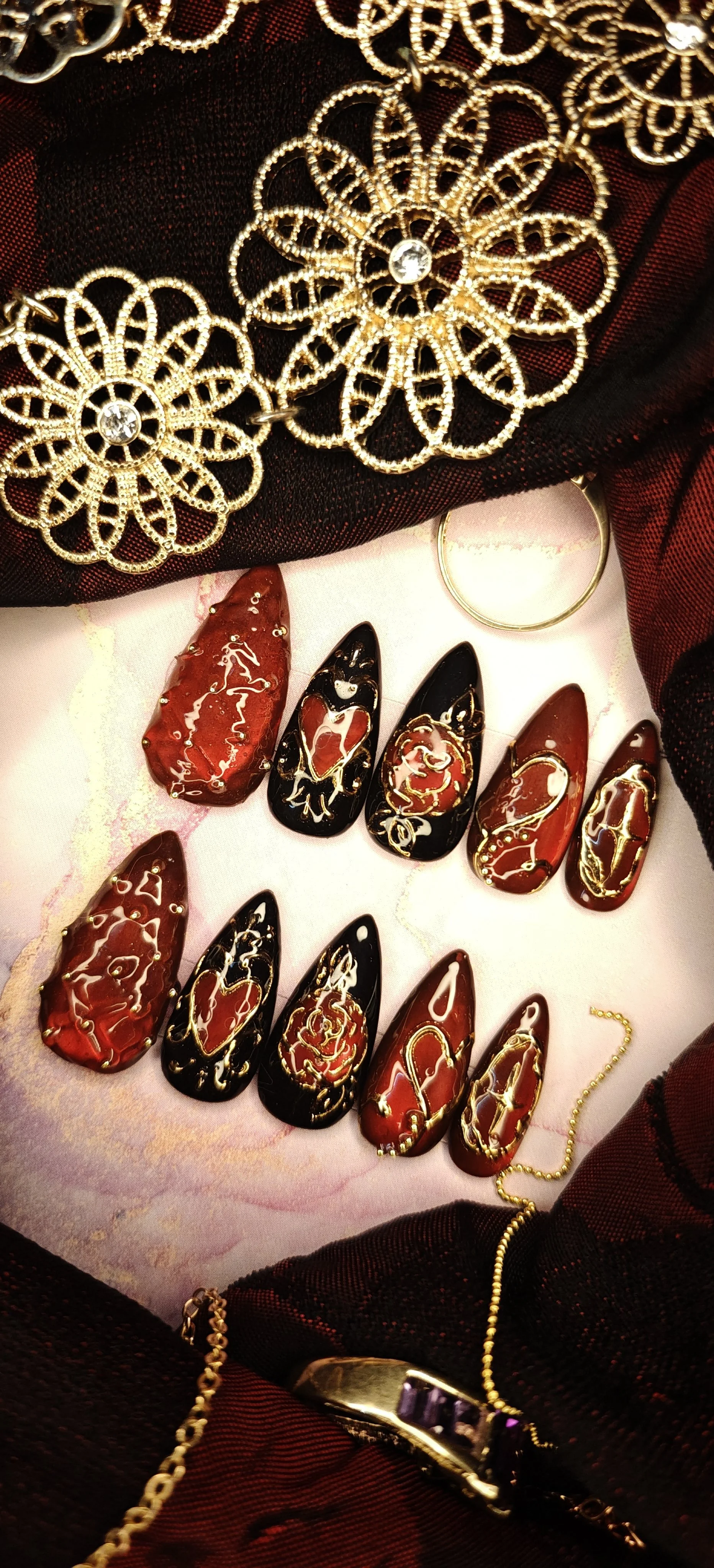 A collection of decorative nail art, jewelry, and accessories displayed on a soft pink surface. The nails are painted red and black with gold accents. Jewelry pieces include gold earrings and rings with embedded stones, along with a gold chain bracelet. A black fabric with gold floral designs surrounds the display.