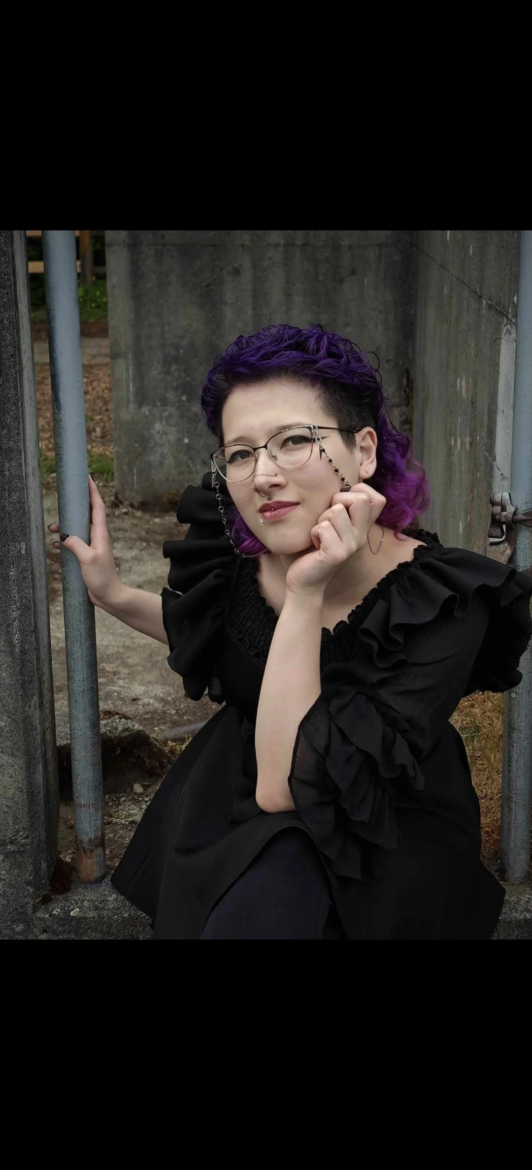 A woman with purple and black hair, wearing glasses and a black ruffled dress, posing outdoors near a concrete structure and metal pipes, holding a chain with glasses.