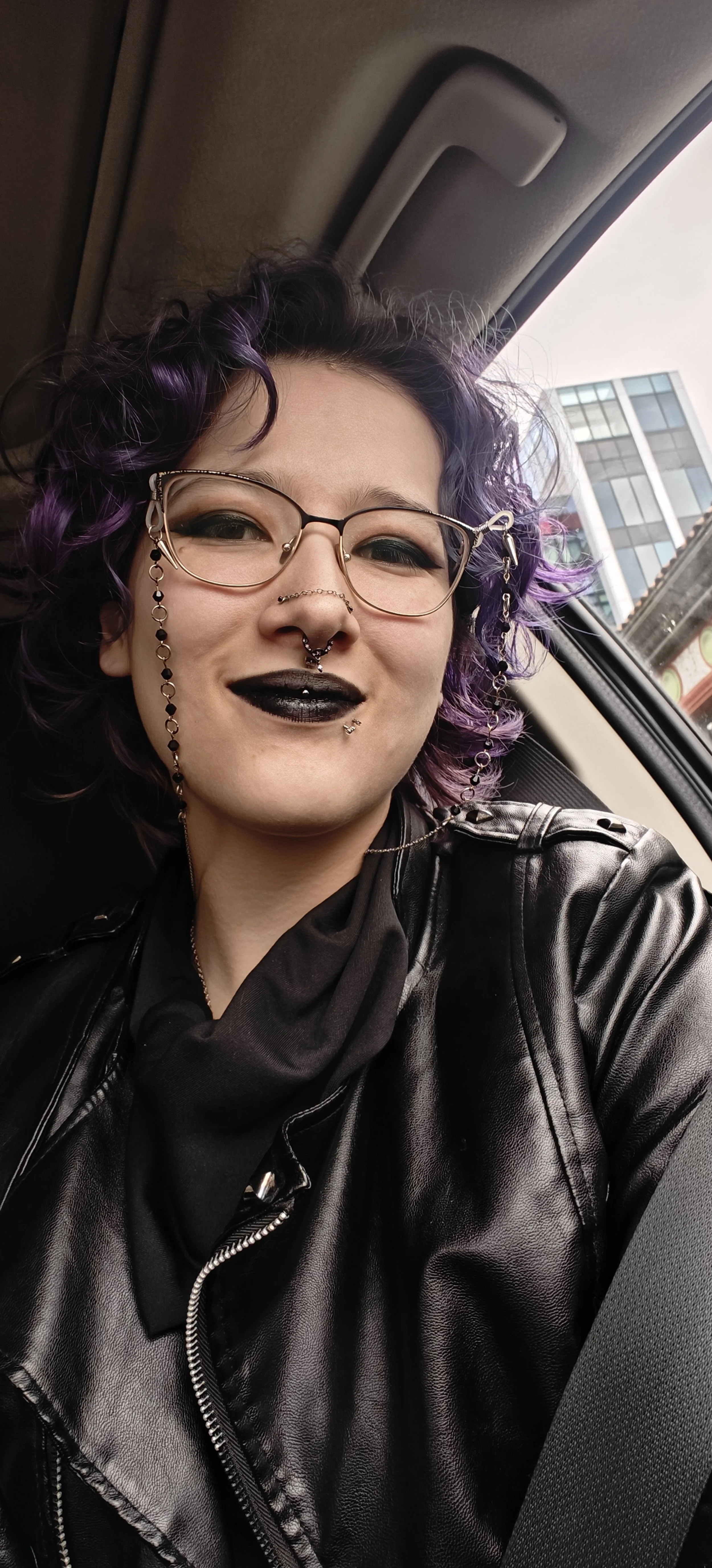 Person with purple and black curly hair, wearing glasses with chain, black lipstick, septum piercing, earrings, leather jacket, sitting in car.