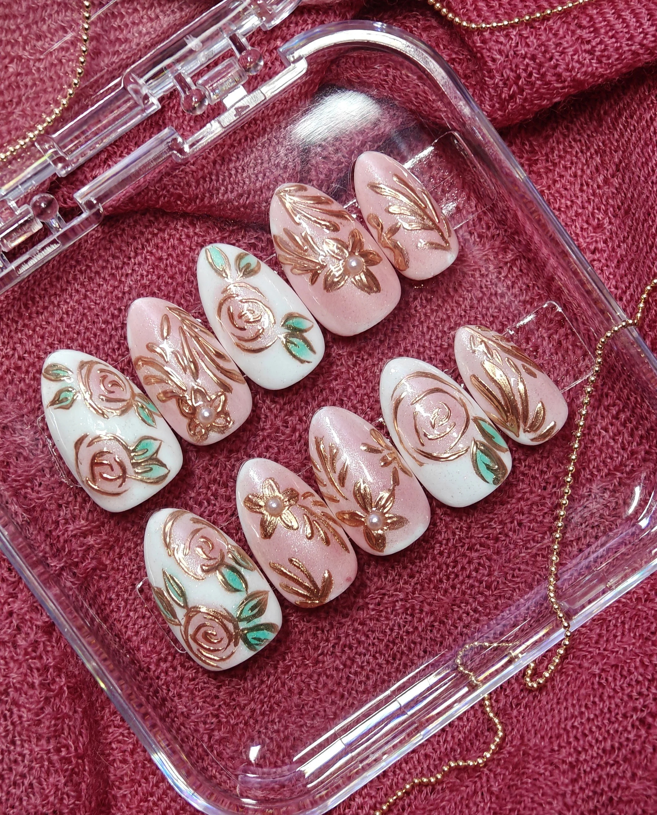 Close-up of decorated false nails with floral and leaf patterns in gold, green, and pink, arranged in a plastic case on a pink textured fabric, with a gold bead necklace partially visible.