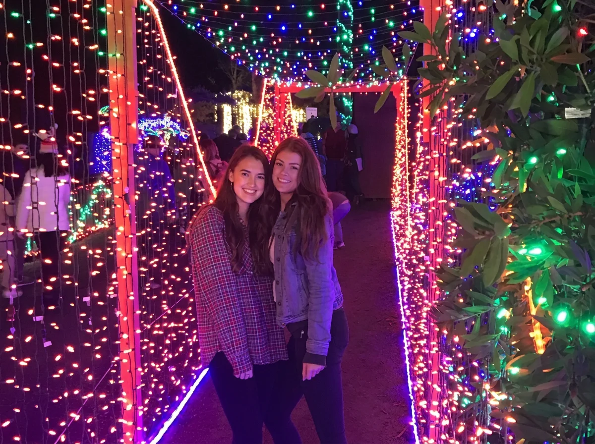 Two young women smiling and posing together at a festive holiday light display with colorful string lights forming an archway, surrounded by illuminated trees and decorations.