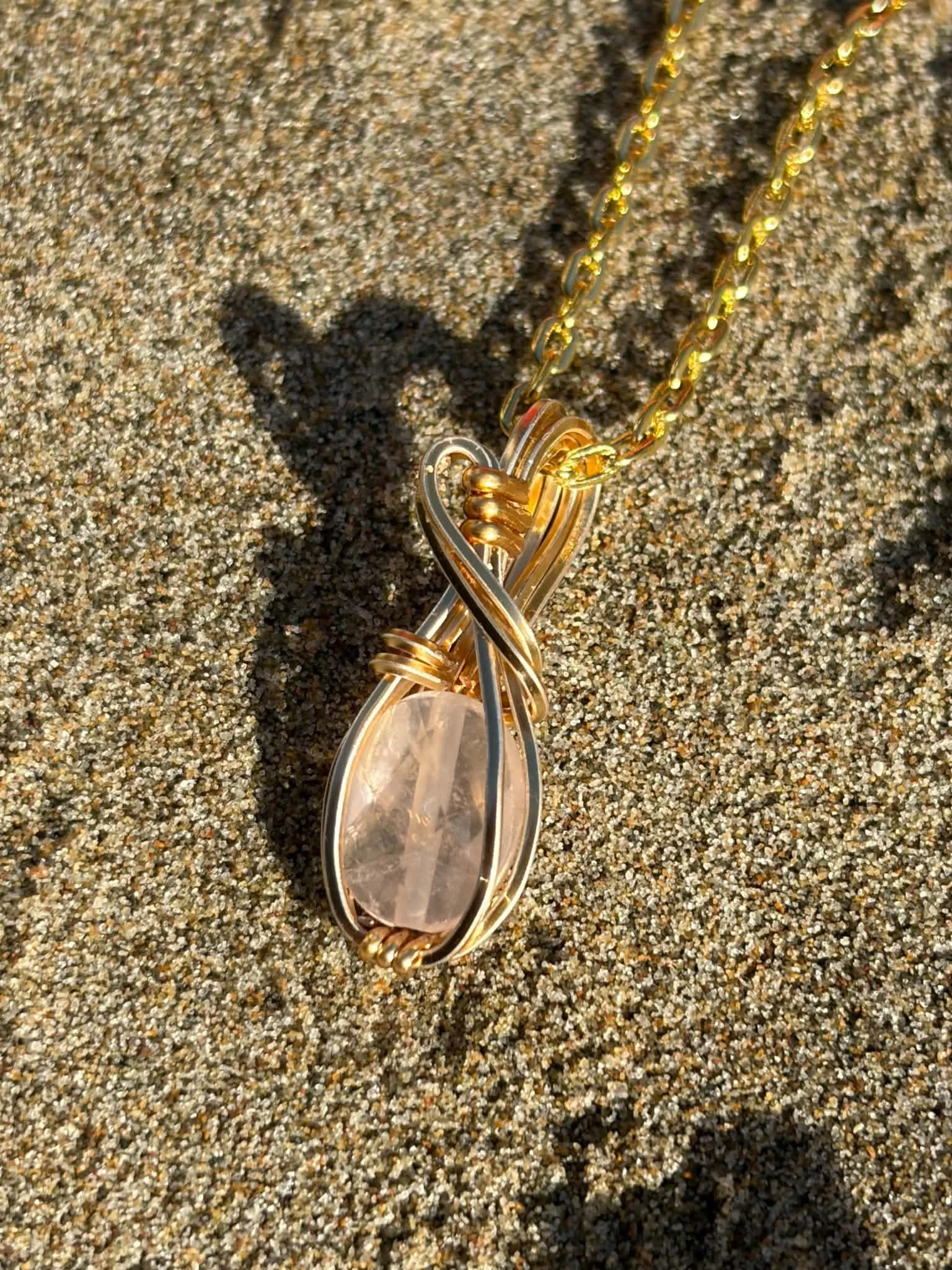 A gold necklace with a pendant lying on sandy ground, casting a shadow.
