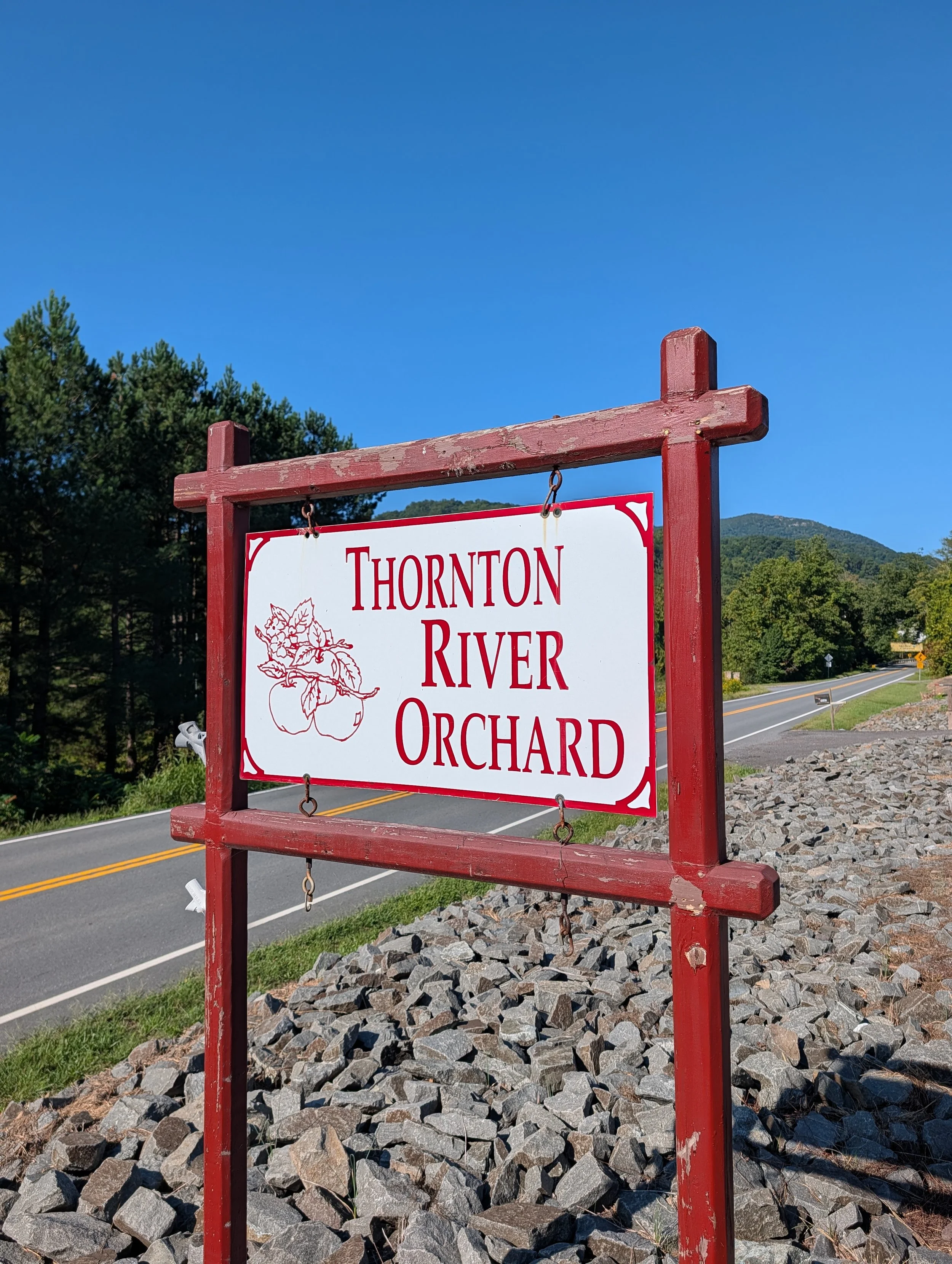 A sign reading 'Thornton River Orchard' in a red wooden frame, with a background of green trees, a mountain, a road, and a clear blue sky.