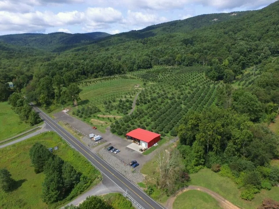 Aerial view of a rural landscape with a red-roofed building, a small parking lot with cars, a two-lane road, and lush green hills and farmland.