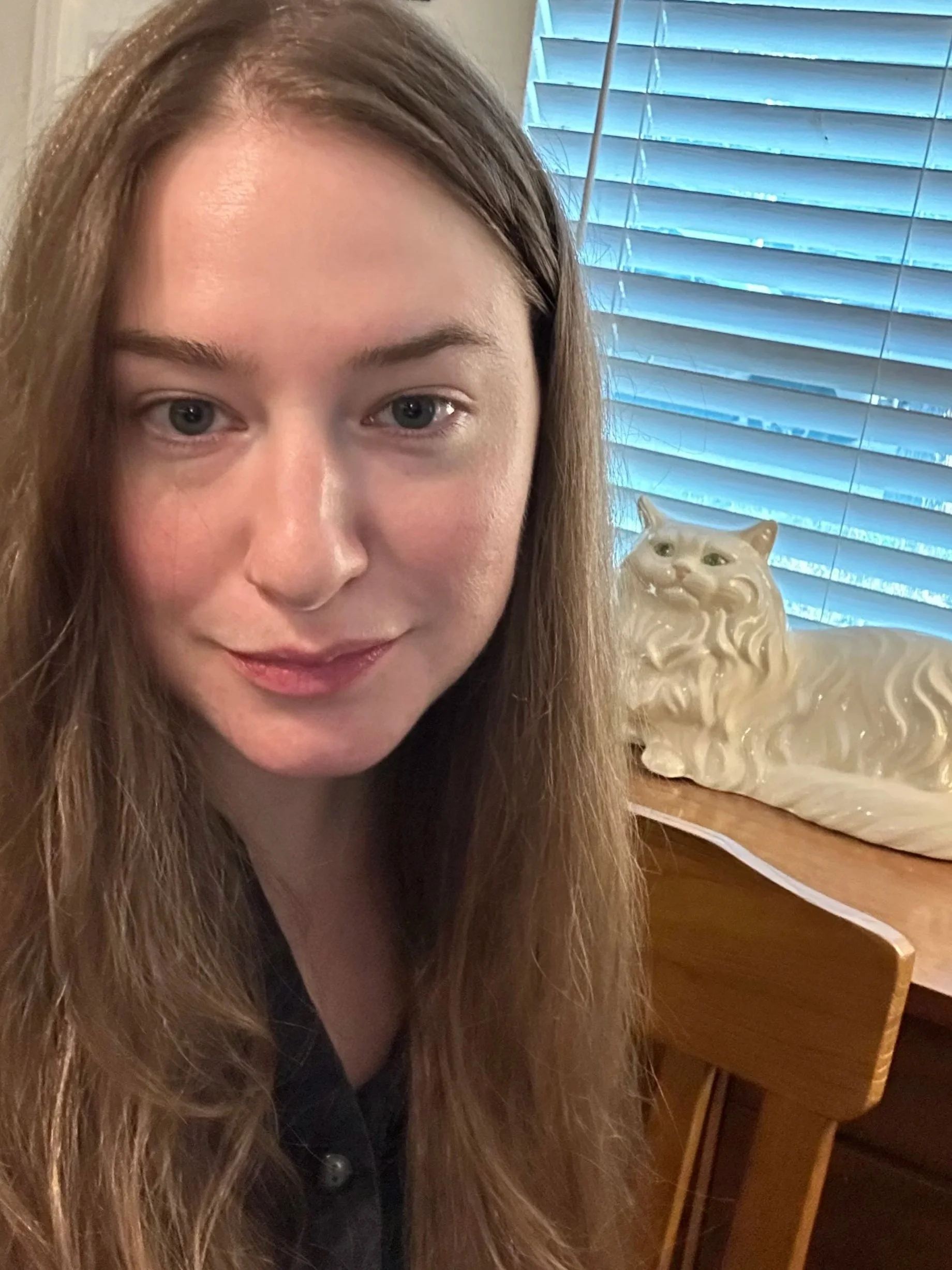 A woman with long, light brown hair and blue eyes is taking a selfie indoors. Behind her is a window with blue blinds and a ceramic sculpture of a lion on a wooden surface.