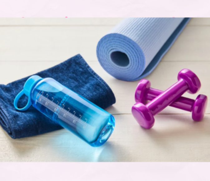 Fitness accessories including a rolled yoga mat, a blue water bottle, a dark towel, and a pair of pink dumbbells on a light wooden surface.