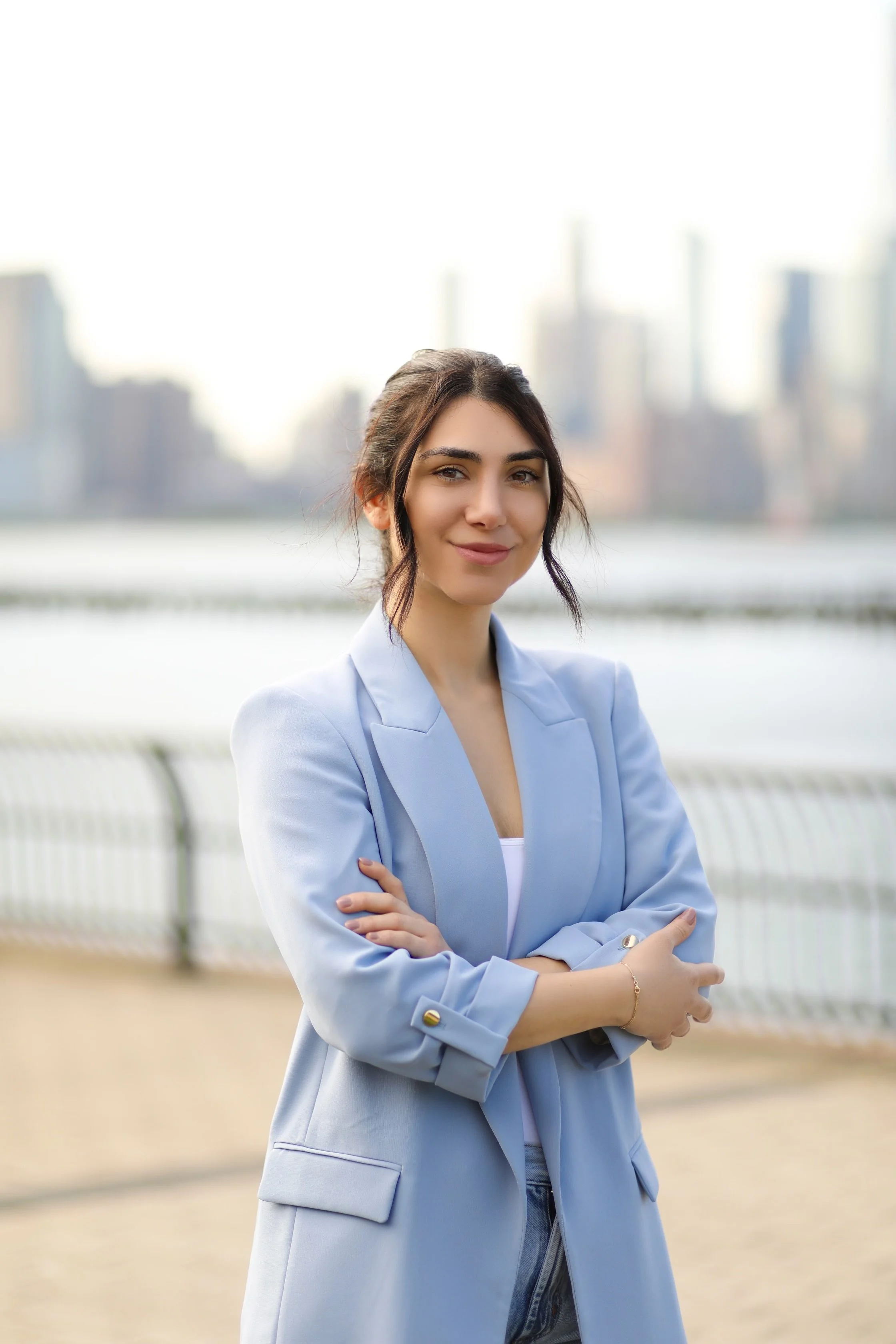 A young woman with dark hair in a bun, wearing a light blue blazer and jeans, standing outdoors near a body of water with a city skyline in the background.