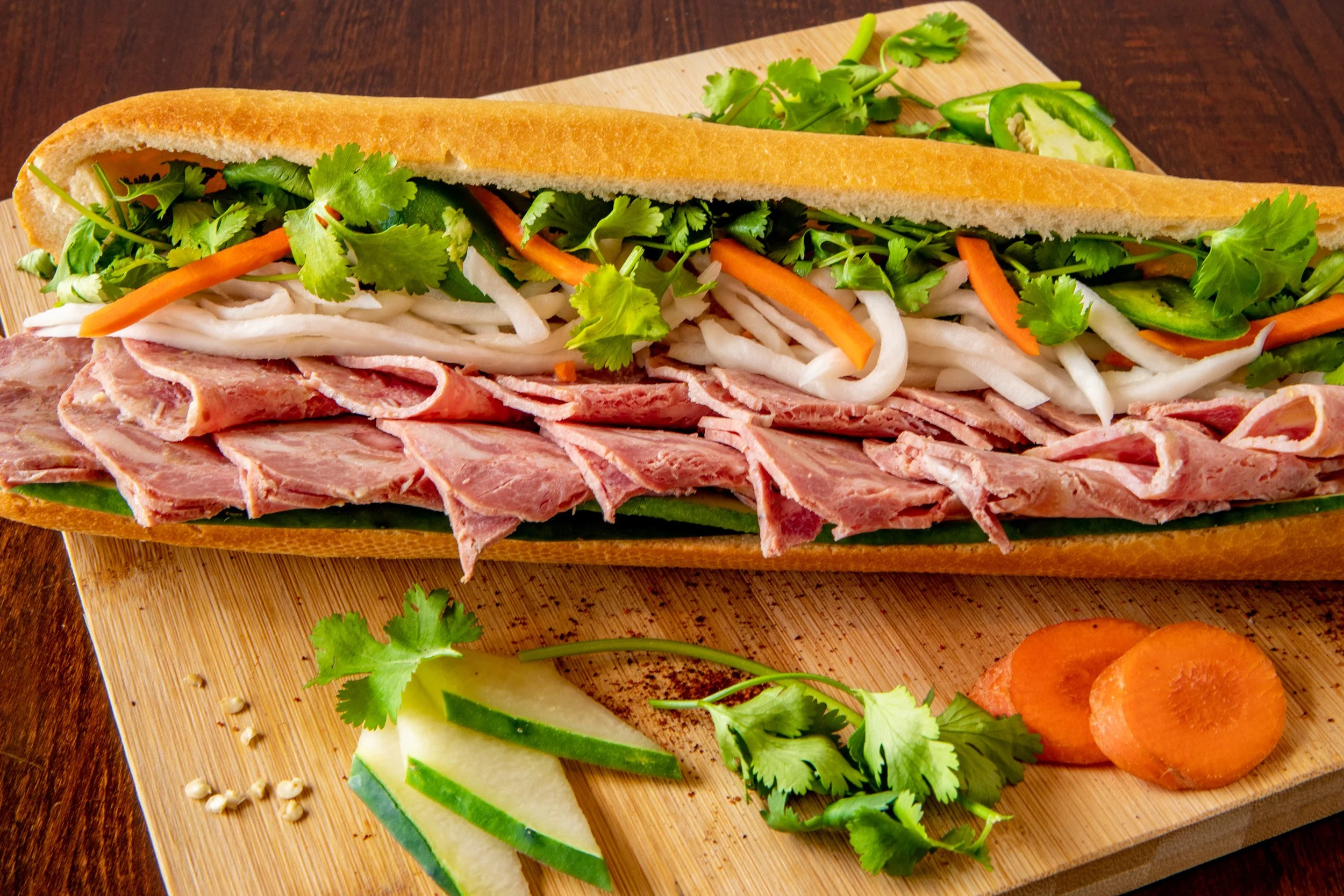A sub sandwich on a wooden cutting board with sliced cucumbers, carrots, cilantro, and shredded lettuce, with a side of orange sauce or ketchup.