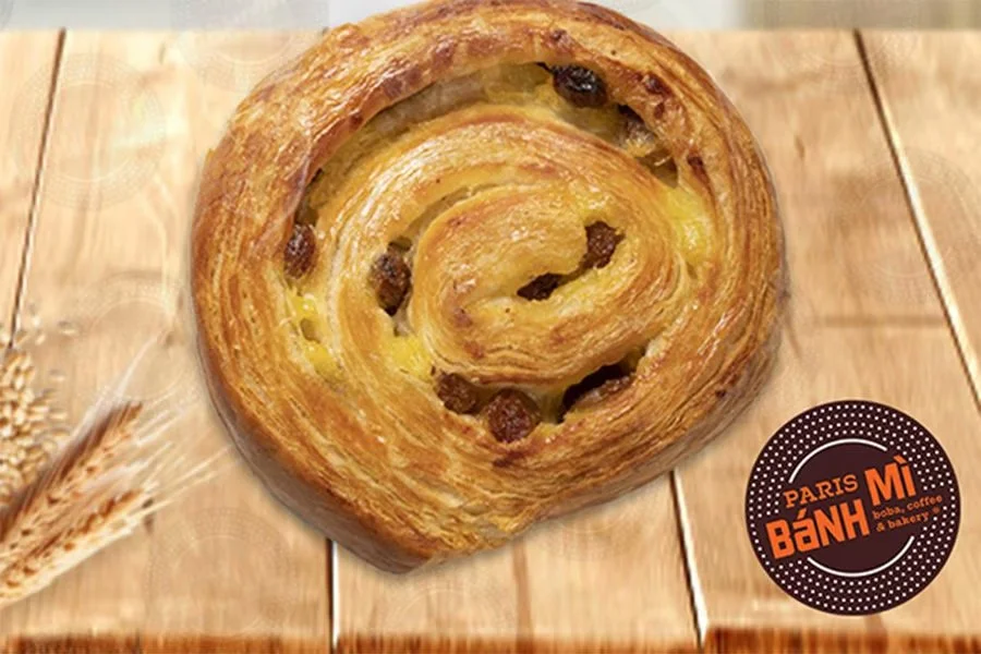 A freshly baked Danish pastry with raisins on a wooden surface. There is a circular logo in the lower right corner that says 'Paris Bánh Mì' in orange and black.