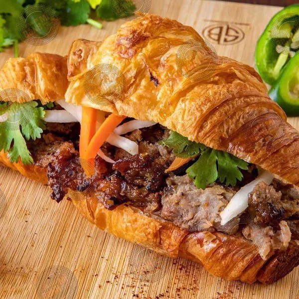Beef sandwich in a croissant bun with cilantro, pickled carrots, and onions on a wooden serving board.