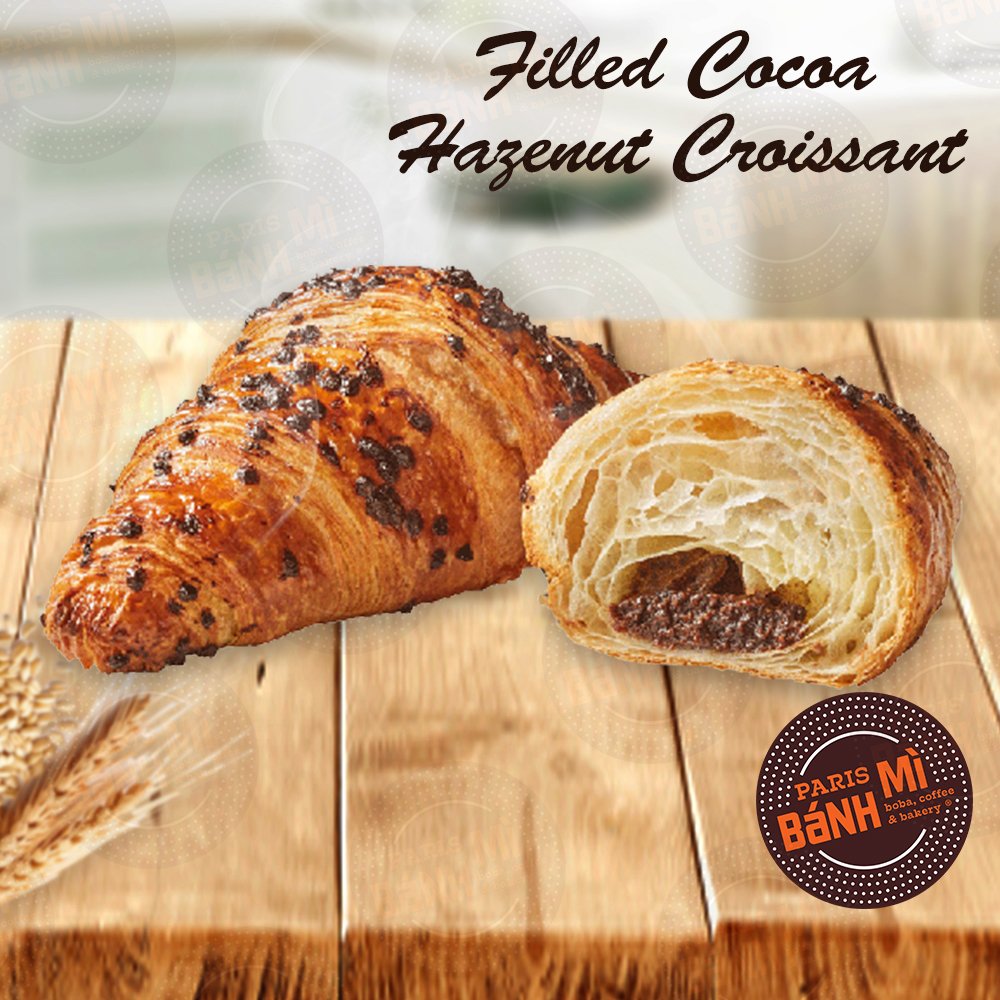 A croissant with filled cocoa hazelnut filling cut in half on a wooden surface. The image includes text indicating it is a filled cocoa hazelnut croissant from Paris Bánh MI, a bakery and coffee shop.
