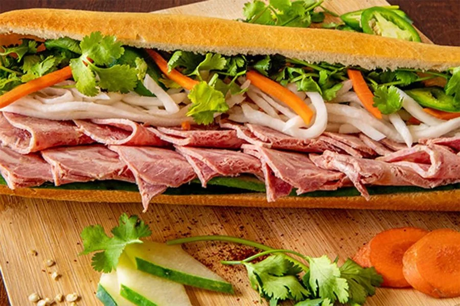 Sub sandwich with sliced roast beef, fresh green herbs, shredded carrots, sliced onions, and cucumber slices on a wooden tray