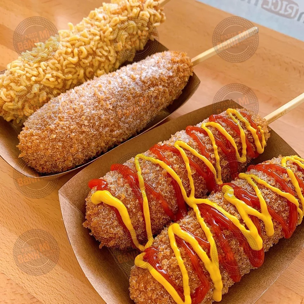 Three corn dogs on sticks, topped with various condiments, on disposable containers. The first has chopped peanuts, the second has a dusting of powdered sugar, and the third has ketchup and mustard drizzle.