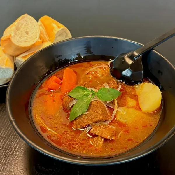 A bowl of beef stew with potatoes, carrots, and herbs, accompanied by slices of bread.