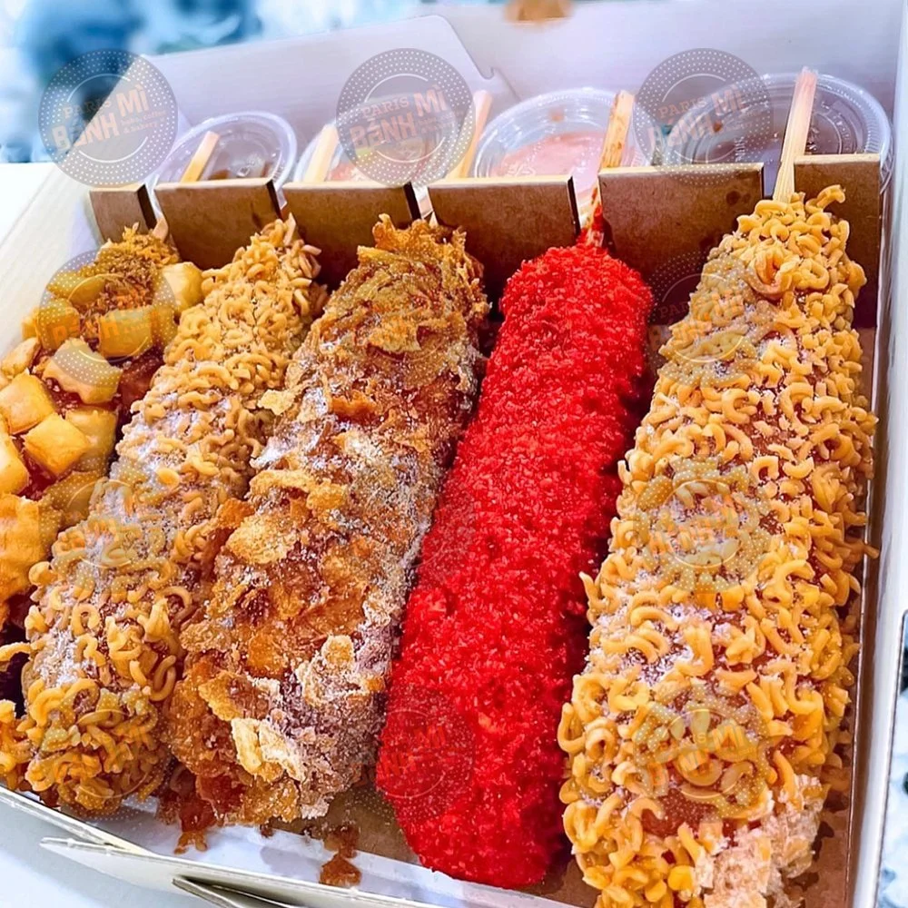 A box of five assorted fried or breaded snack items with dipping sauces in small cups in the background.