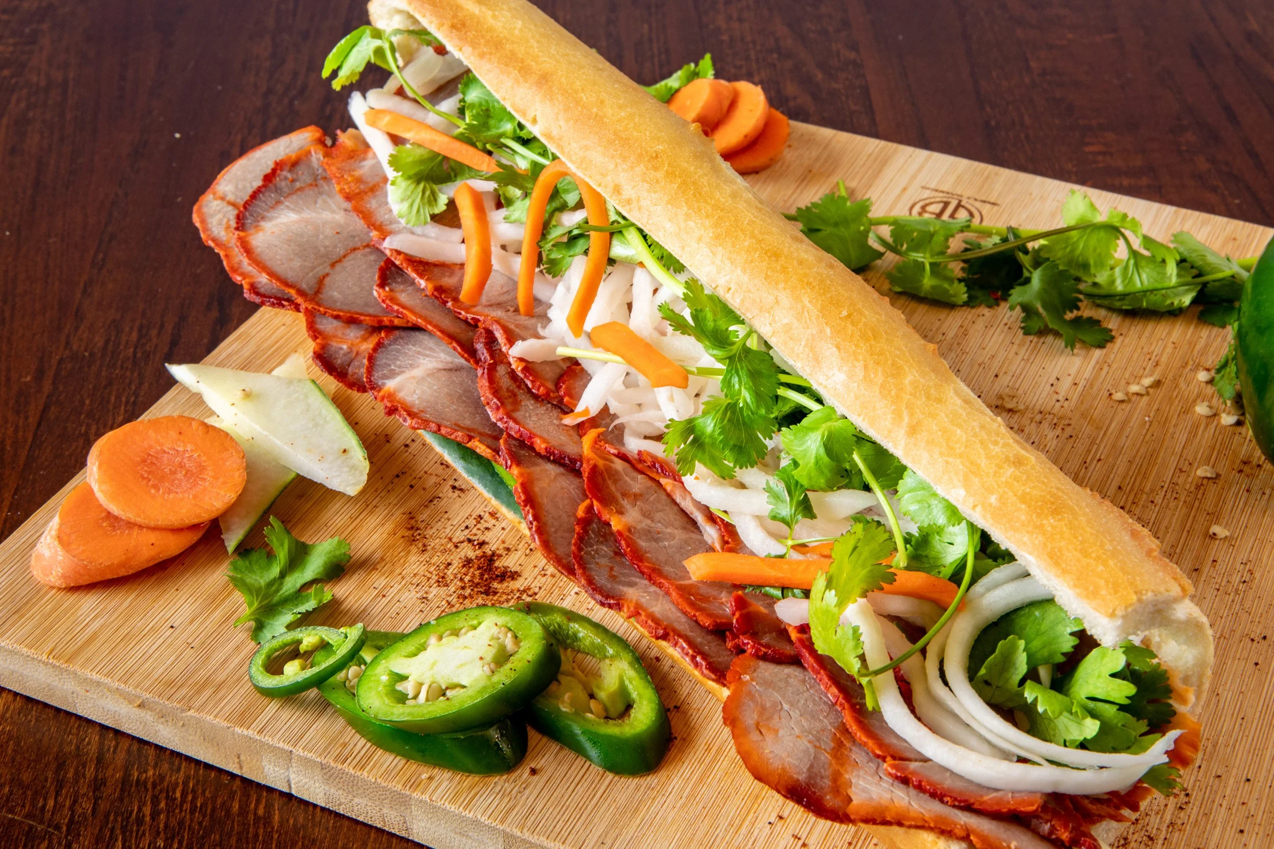 A fresh bánh mì sandwich with sliced pork, pickled vegetables, cilantro, and jalapeño peppers on a wooden cutting board, with sliced carrots, cucumbers, and cilantro around it.