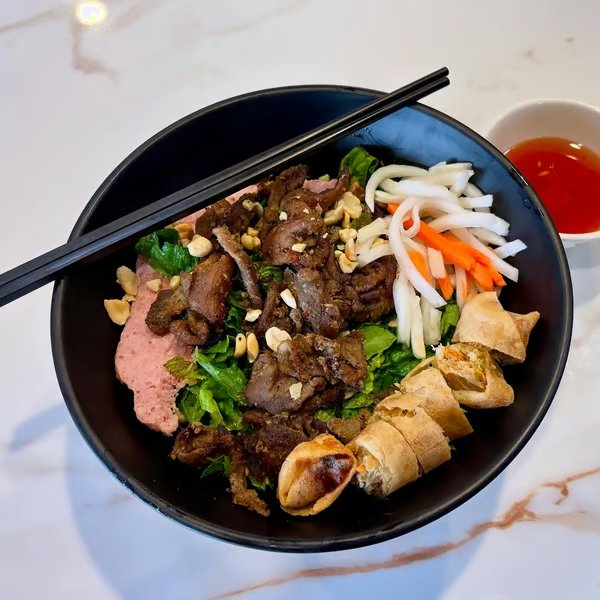 A bowl of Vietnamese bun thit nuong noodles with grilled meat, fresh vegetables, spring rolls, crushed peanuts, and a side of dipping sauce.