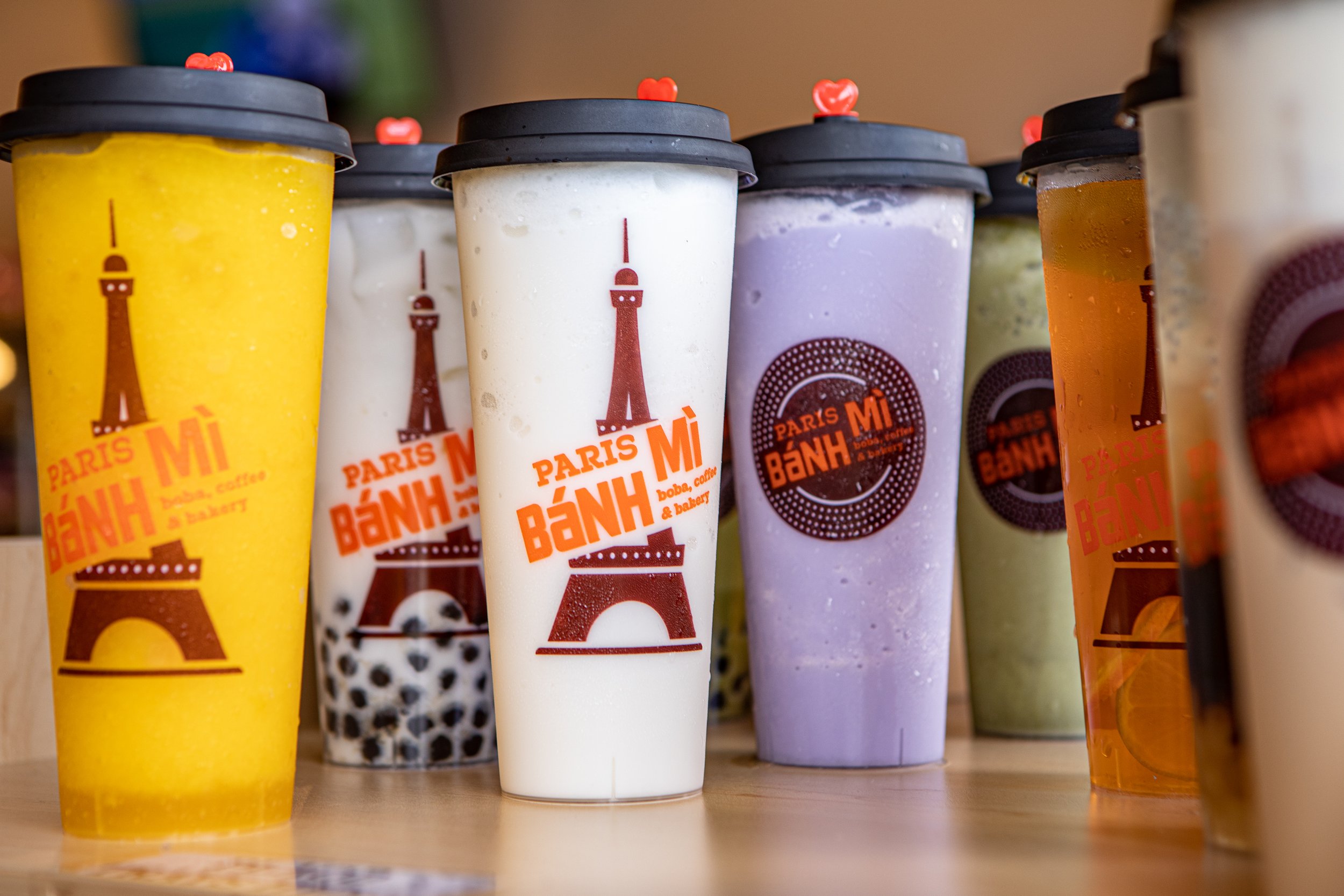 A row of colorful smoothie cups with black lids, decorated with small heart toppers, and featuring Paris-themed stickers, including the Eiffel Tower.