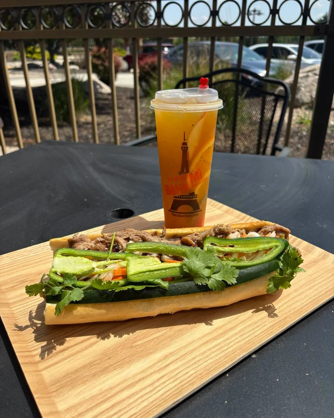 A banh mi sandwich with sliced cucumbers, carrots, cilantro, and beef on a hoagie roll, accompanied by a tall iced tea in a plastic cup with a straw, on a wooden serving board outdoors.