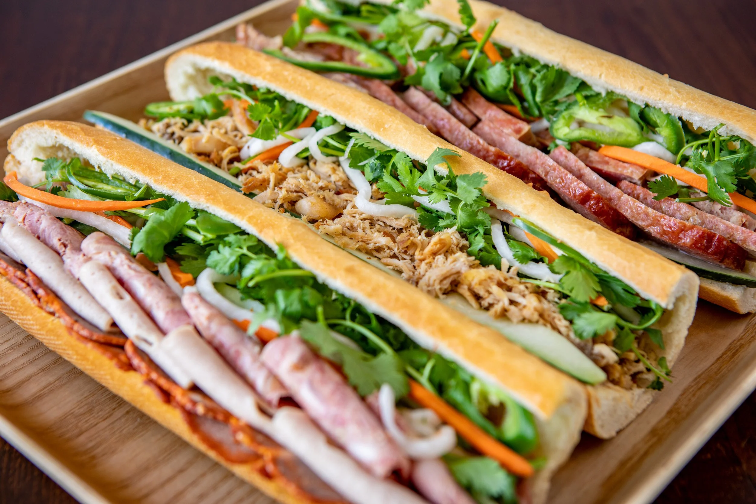 Close-up of a tray of bánh mì sandwiches filled with fresh vegetables, herbs, and sliced meats on baguette buns.