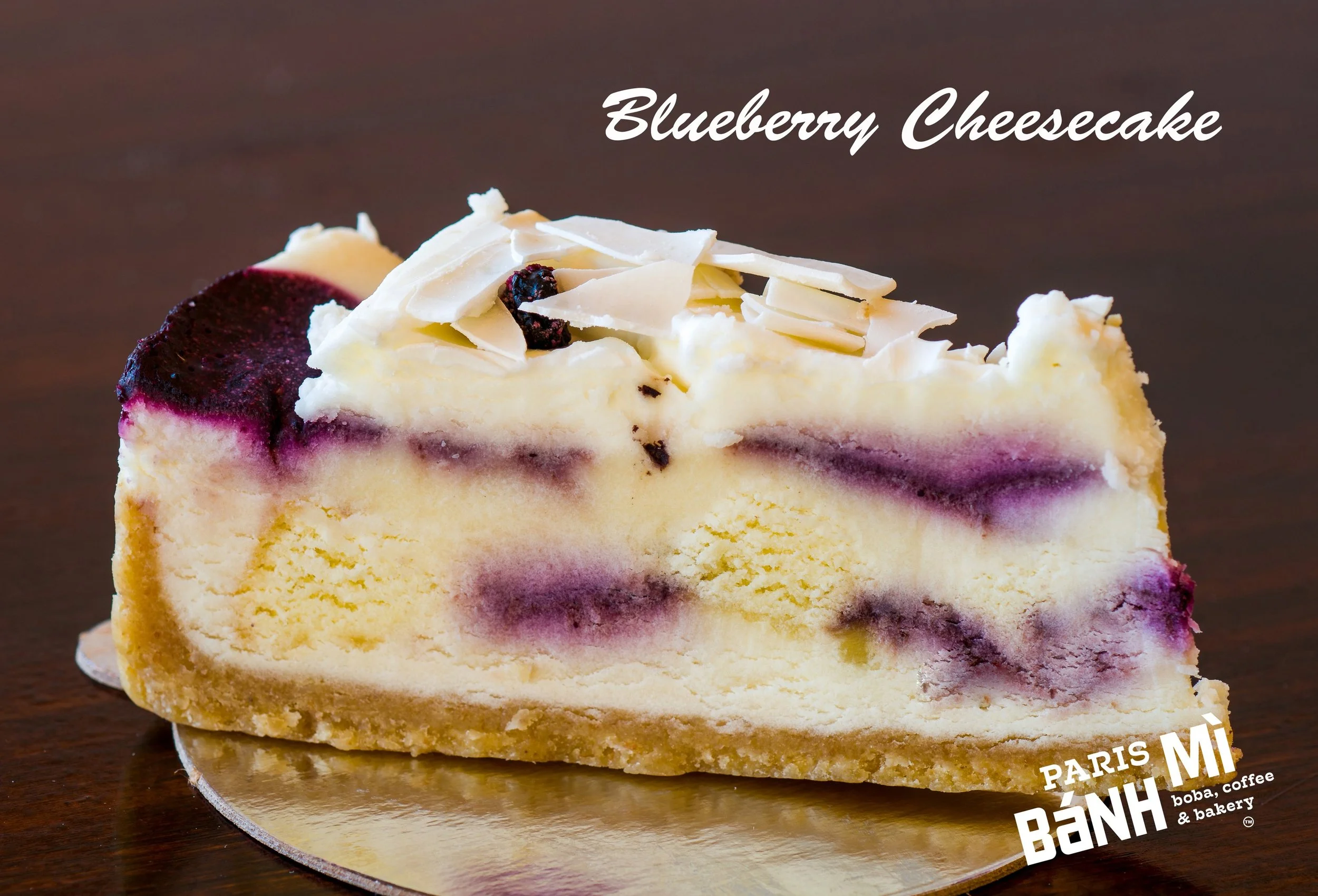A slice of blueberry cheesecake with white chocolate shavings on top and a dark wooden surface in the background.