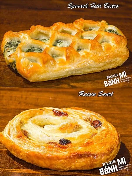 Two baked pastry dishes on a wooden surface. The top pastry is a spinach feta bistro, rectangular with a golden crust and toppings of spinach and feta cheese. The bottom pastry is a raisin swirl, round with a flaky crust and embedded raisins.