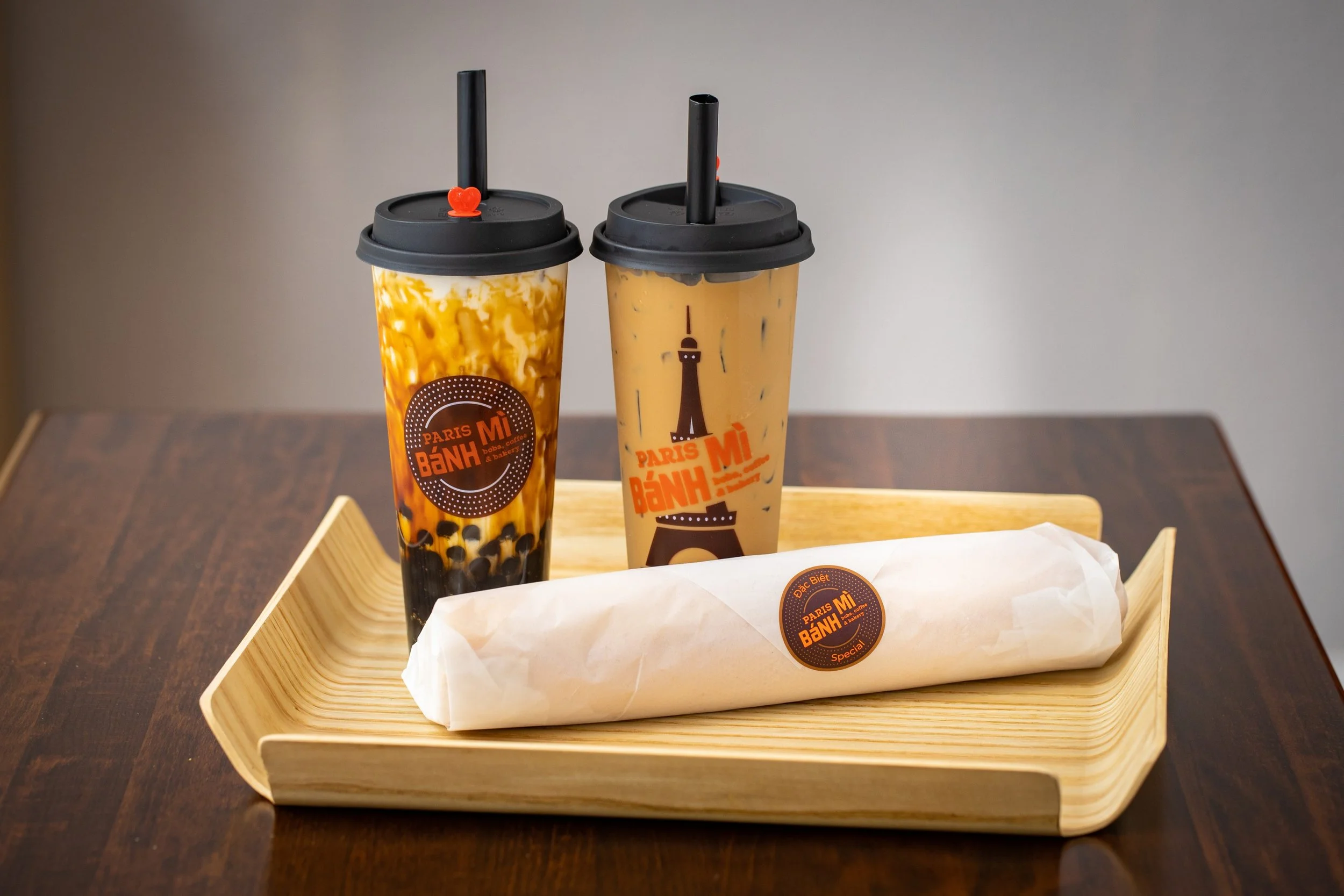 Two plastic cups of bubble tea with black straws, placed on a wooden tray on a dark wooden table.