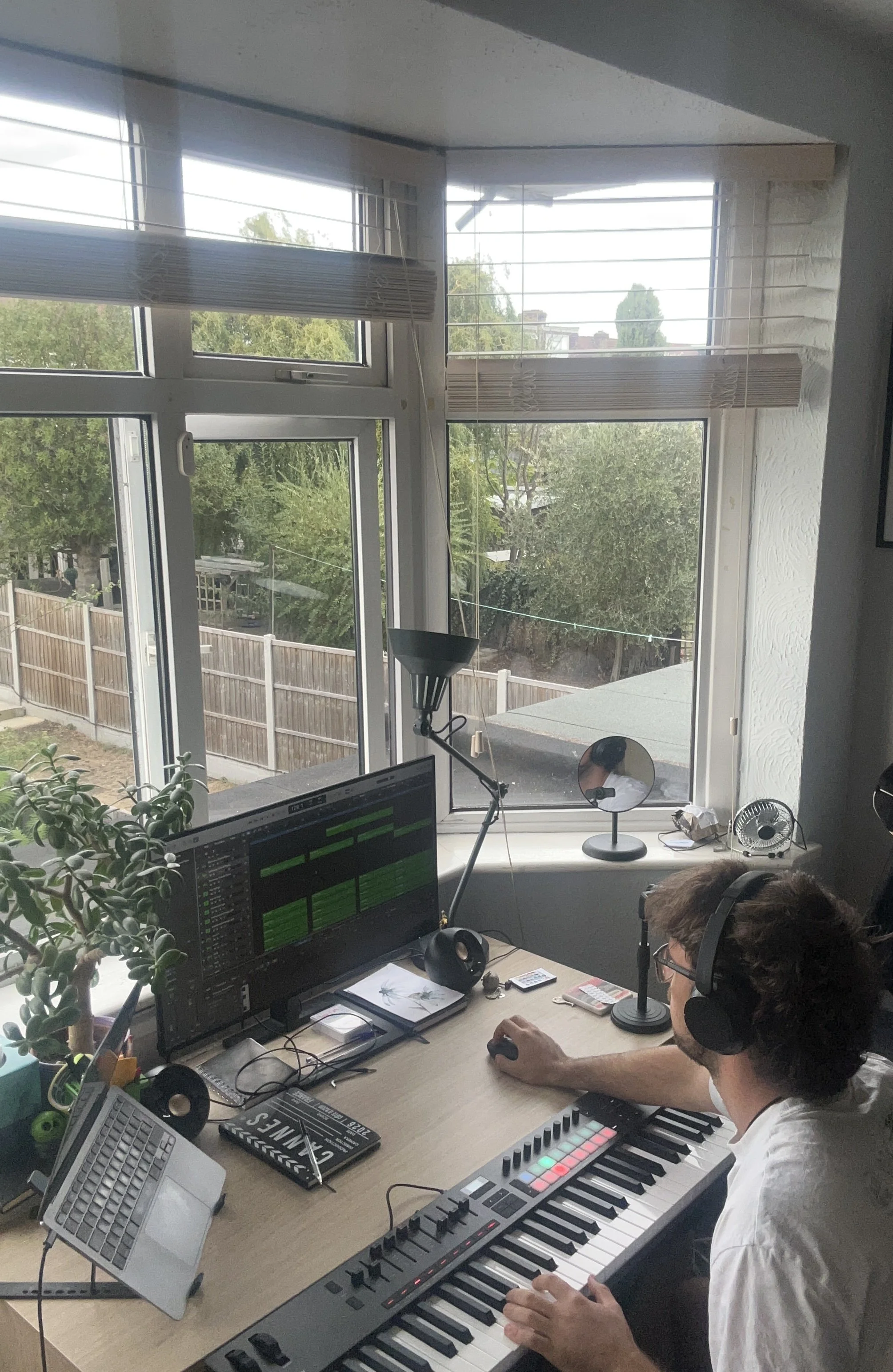 A person wearing glasses and headphones sitting at a desk, using a MIDI keyboard controller, with an Apple monitor, a small electronic keyboard, and other electronic music production equipment inside a room with large windows and a view of trees outdoors.