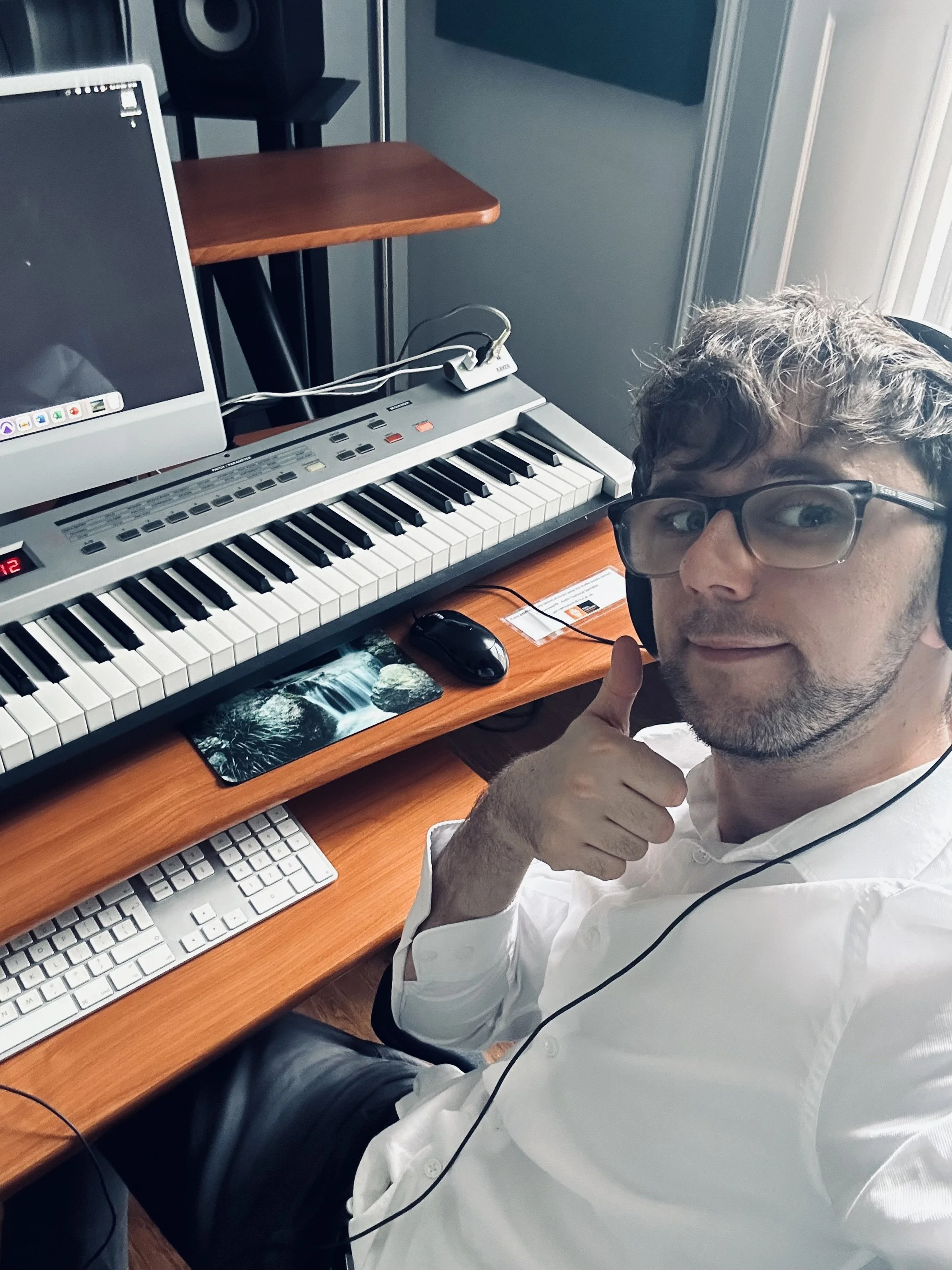 A person with glasses and a white shirt, sitting at a desk with a computer and keyboard, giving a thumbs-up gesture in a home office environment.