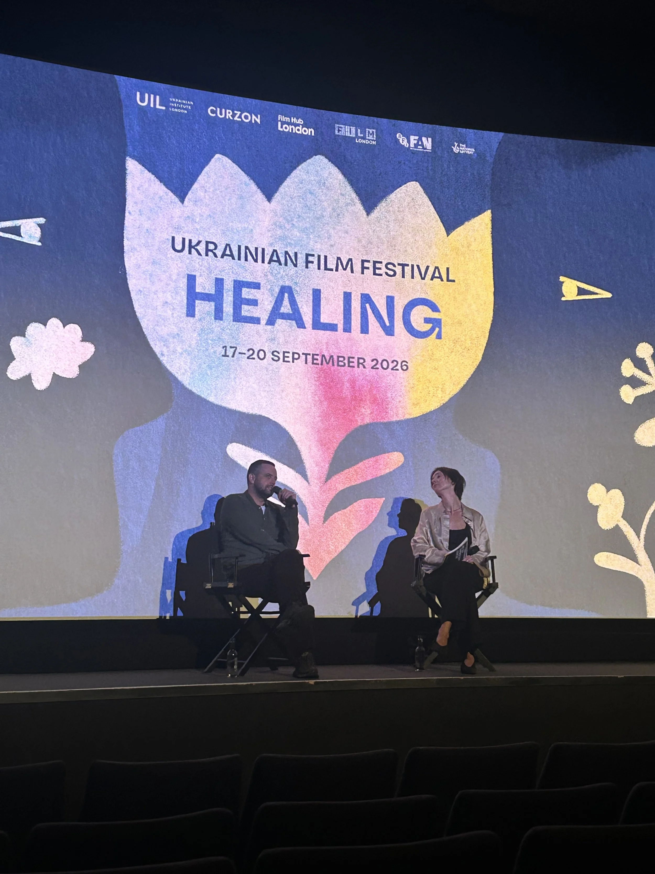 A stage at the Ukrainian Film Festival Healing in September 2026 with two panel speakers seated and a large screen displaying the event's branding, including logos and event dates.
