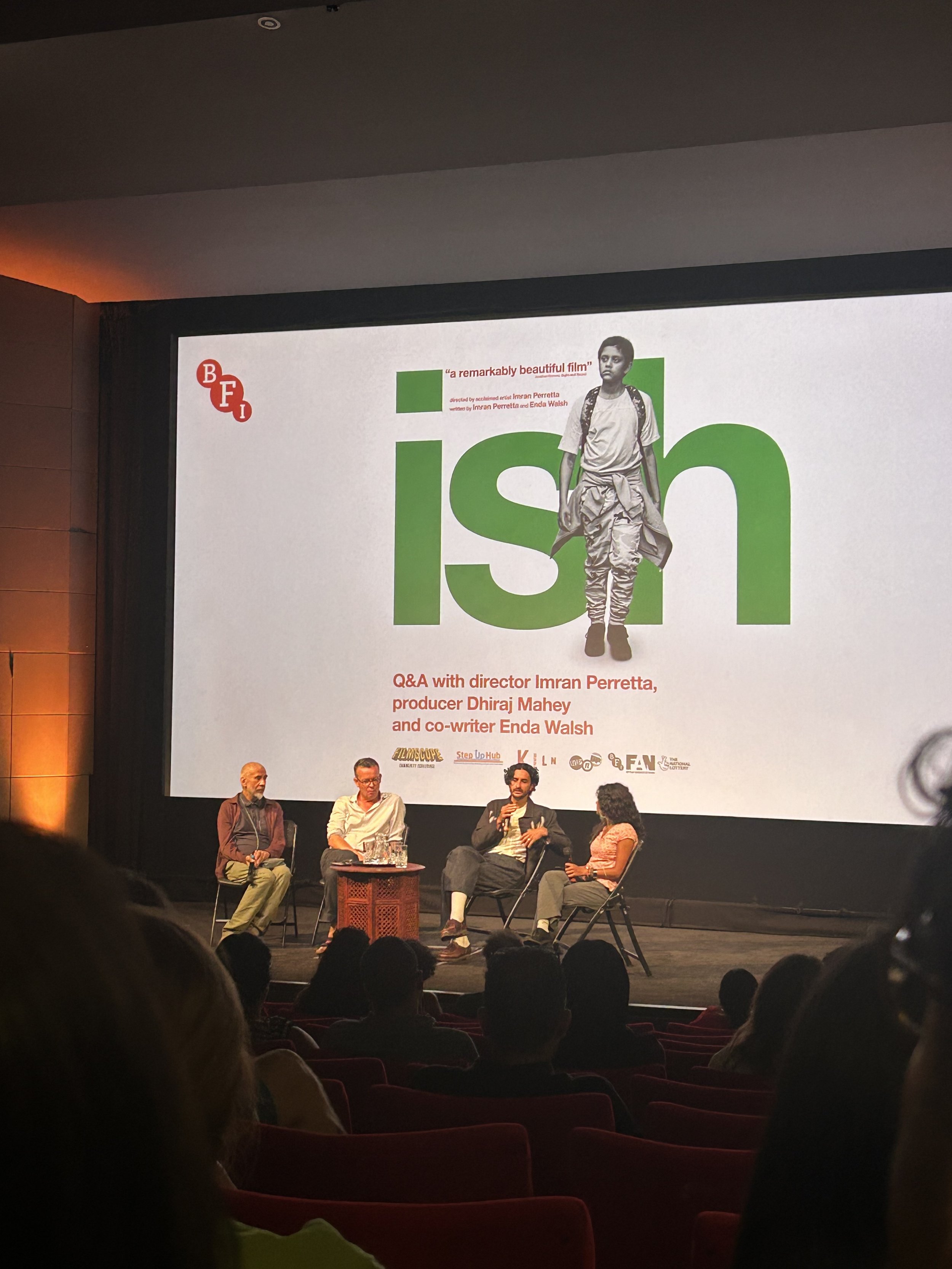 A panel discussion on stage with four people seated and speaking into microphones, in front of a large screen displaying promotional material for a film titled 'Ish', with a young boy dressed in casual clothing and a backpack, and audience members visible in the foreground.