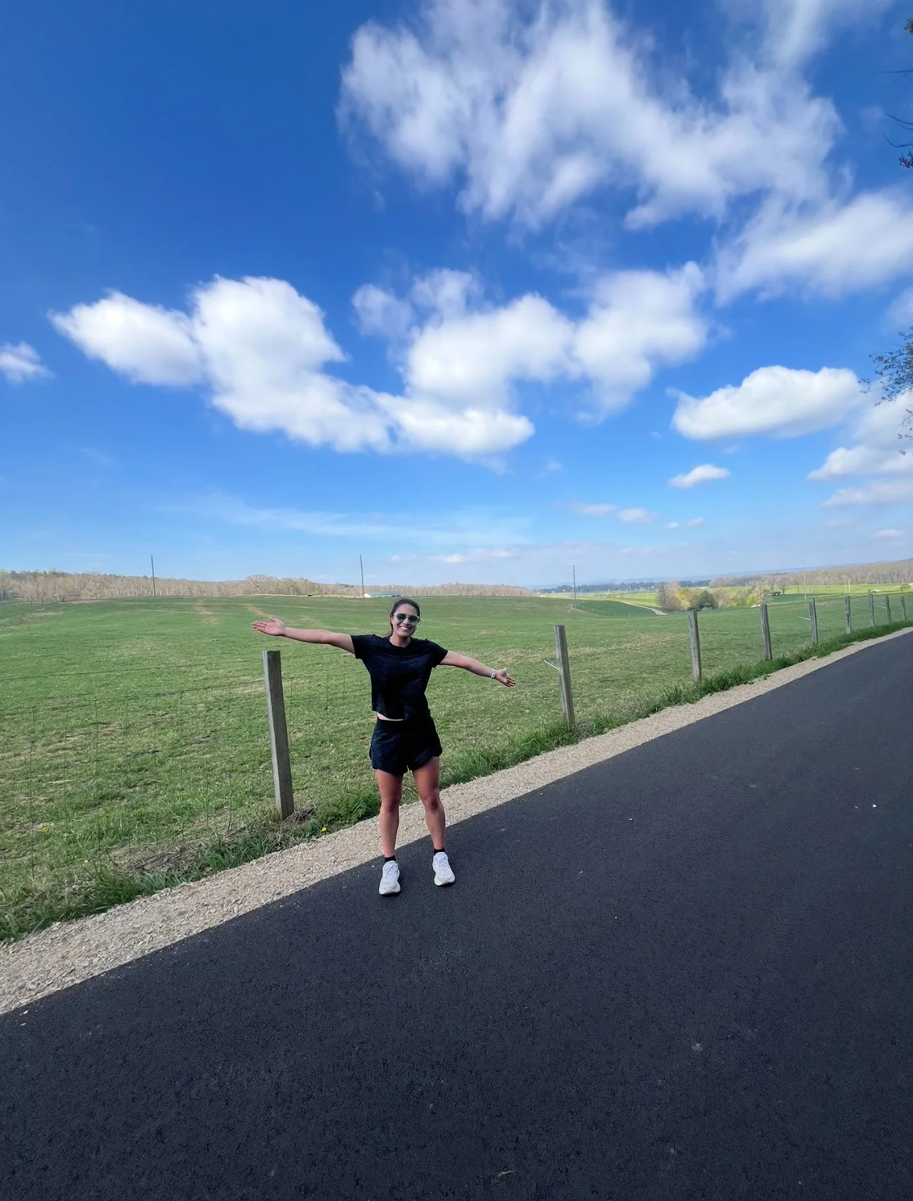 A woman smiling with arms outstretched standing on the side of a paved road, with a fence and a green field stretching into the distance. The sky above is blue with scattered white clouds.